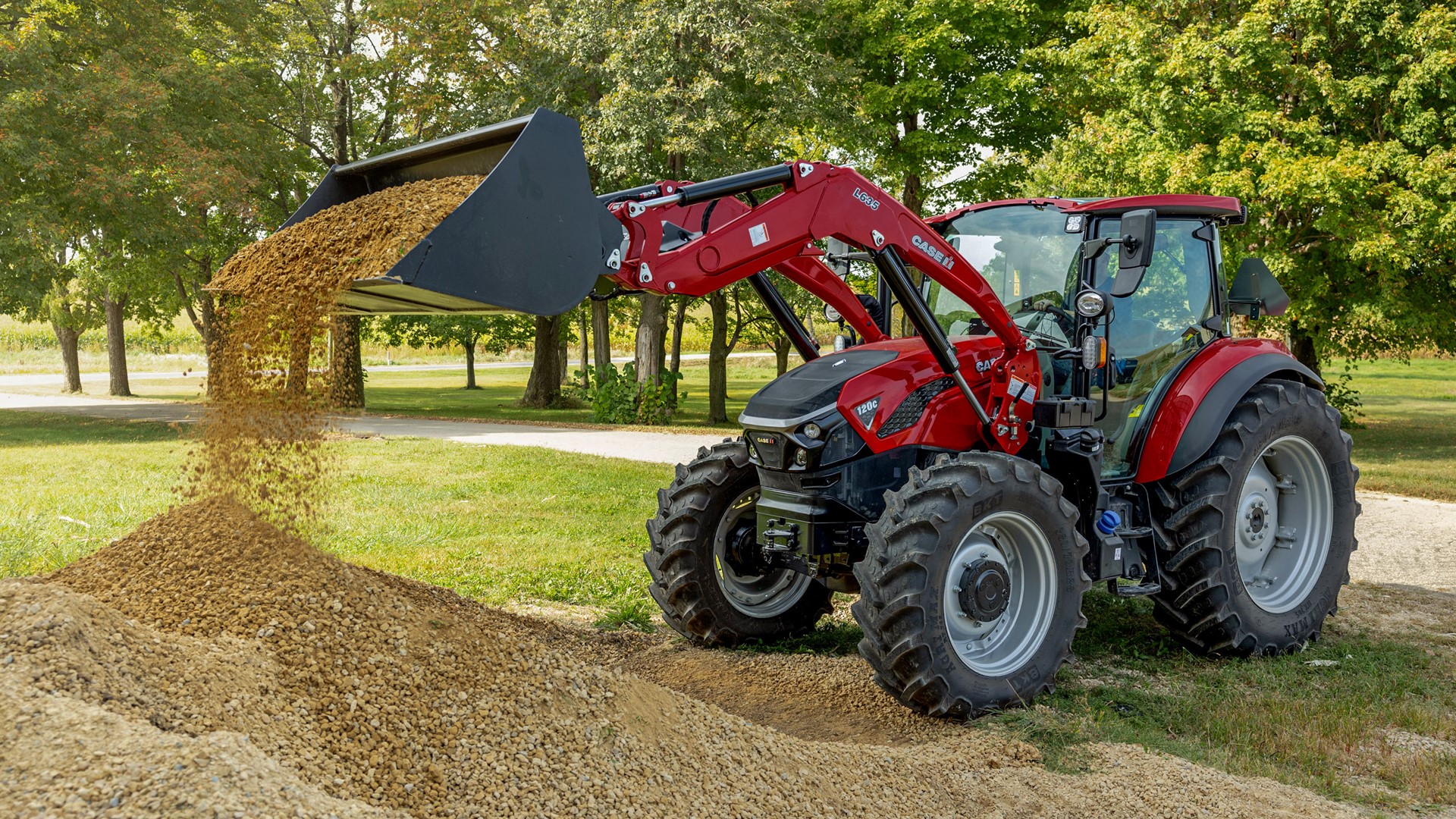 A red tractor with a front loader is dumping a pile of gravel onto the ground in a green outdoor setting. The tractor is positioned on a dirt path surrounded by trees, showcasing a sunny day. The loader is raised, and gravel is cascading down from the bucket.