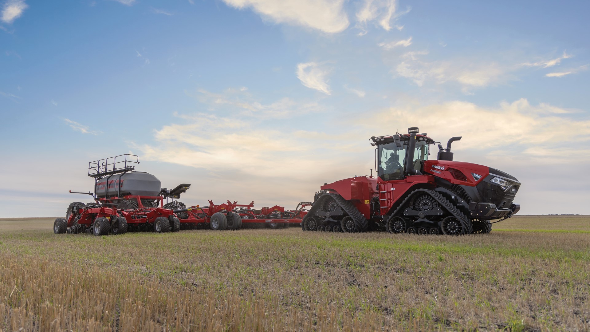 A large red Case IH tractor with rubber tracks is pulling a wide agricultural planter across a field under a partly cloudy sky during sunset or sunrise. The field has short stubble and green patches, indicating recent harvesting or planting activity.