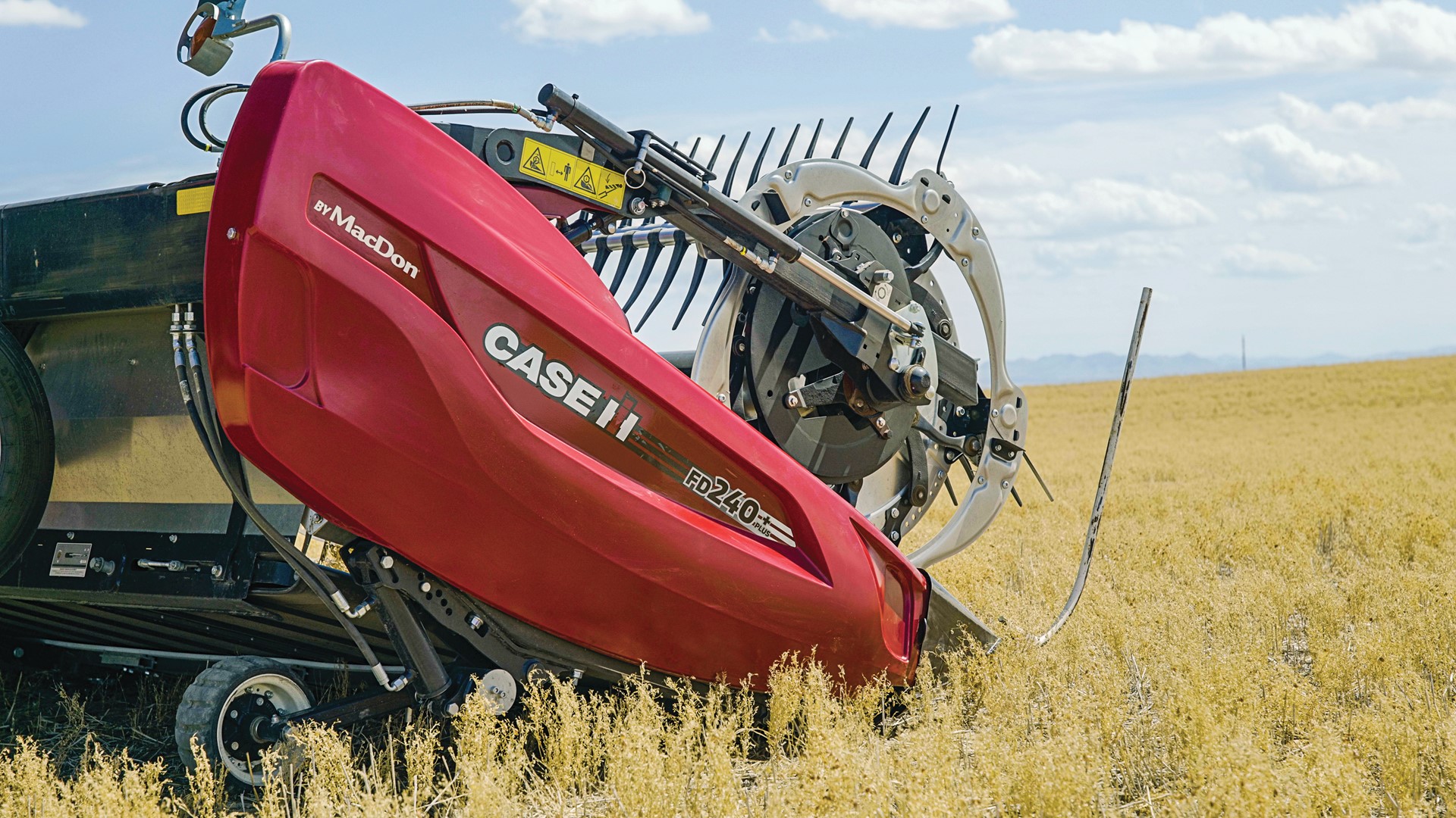 A close-up view of a red Case IH FD240 agricultural harvesting machine by MacDon operating in a golden crop field under a partly cloudy blue sky. The machine's cutting and gathering components are visible, with dry crops surrounding it.