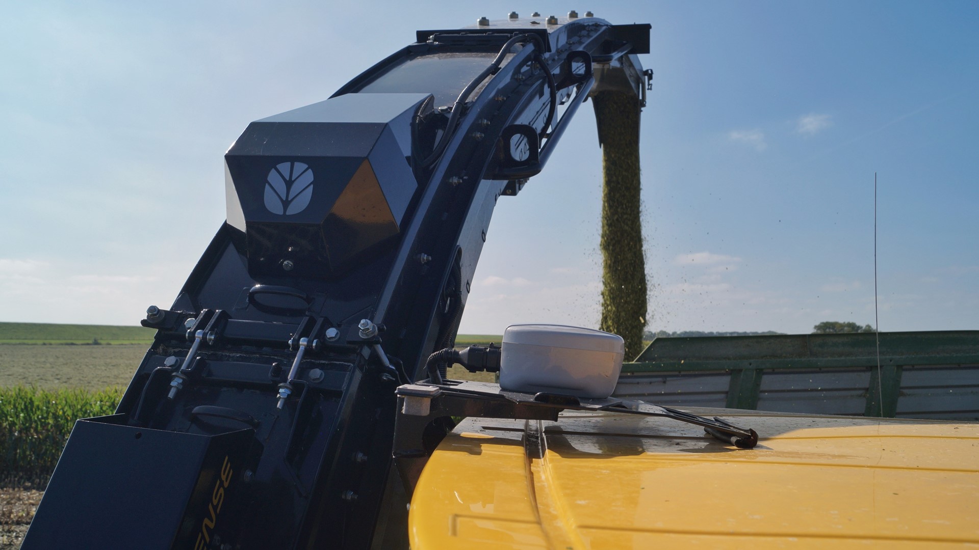 Close-up of a modern agricultural harvester unloading freshly harvested crops through a chute into a trailer on a sunny day with clear blue sky and green fields in the background.