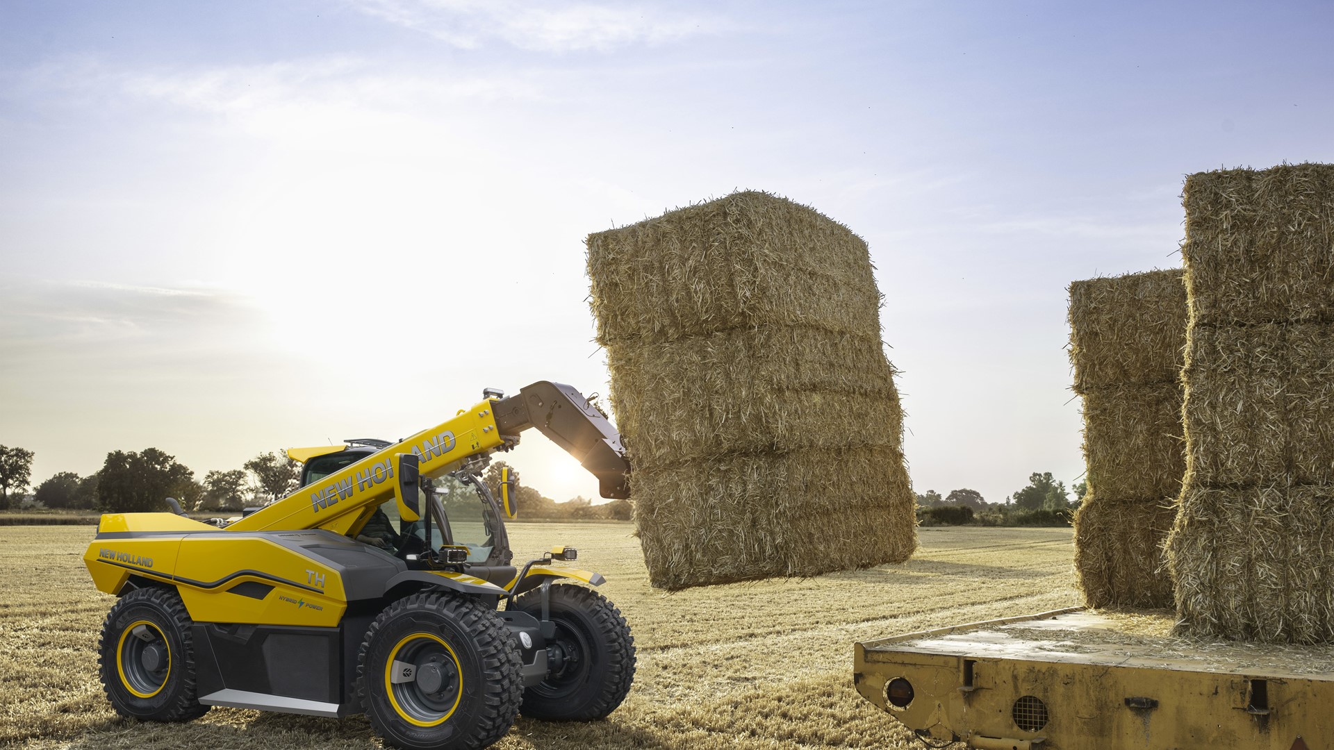 A yellow New Holland telehandler lifting large rectangular hay bales in a harvested field during sunset, with a flatbed trailer nearby stacked with more hay bales.