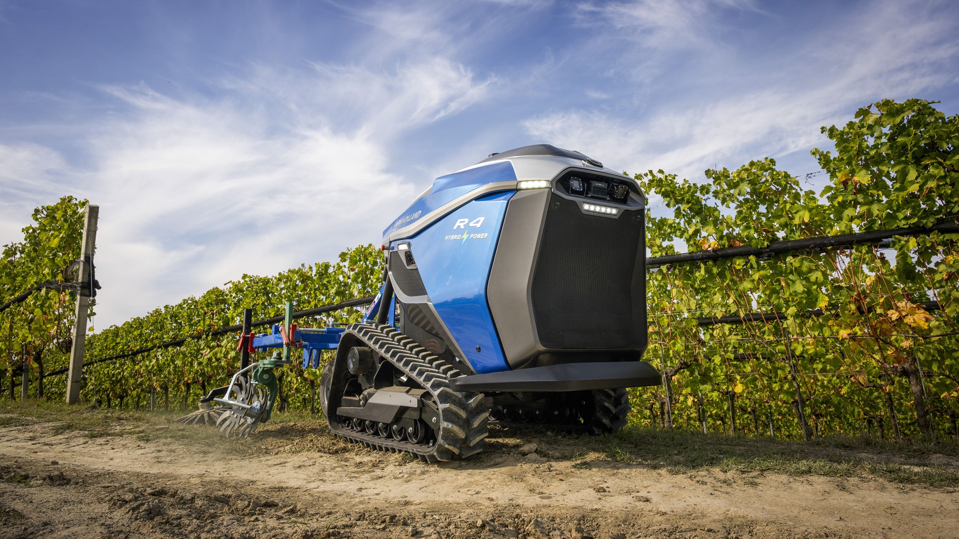 A blue autonomous agricultural robot with tank-like tracks working in a vineyard under a partly cloudy sky, equipped with farming tools for soil cultivation.