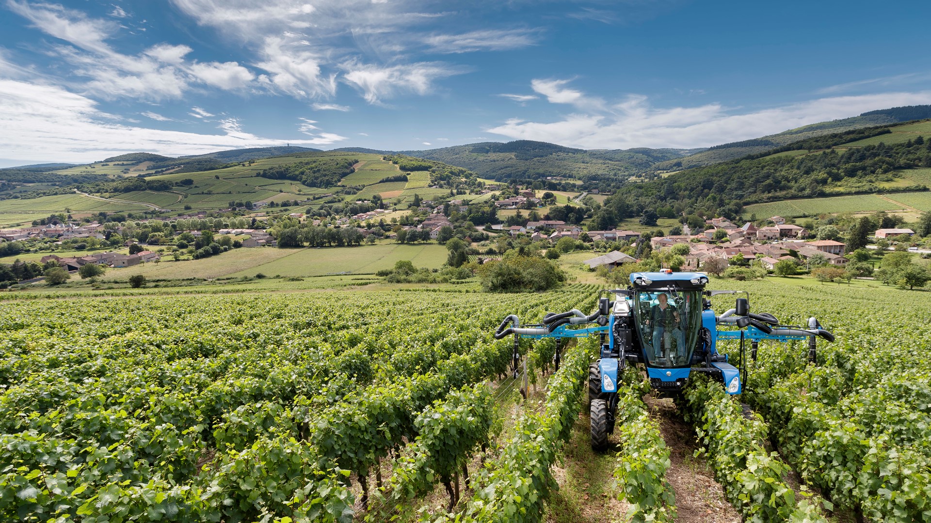 A blue agricultural tractor working in a lush green vineyard with rows of grapevines under a partly cloudy blue sky, with rolling hills and a small village in the background.
