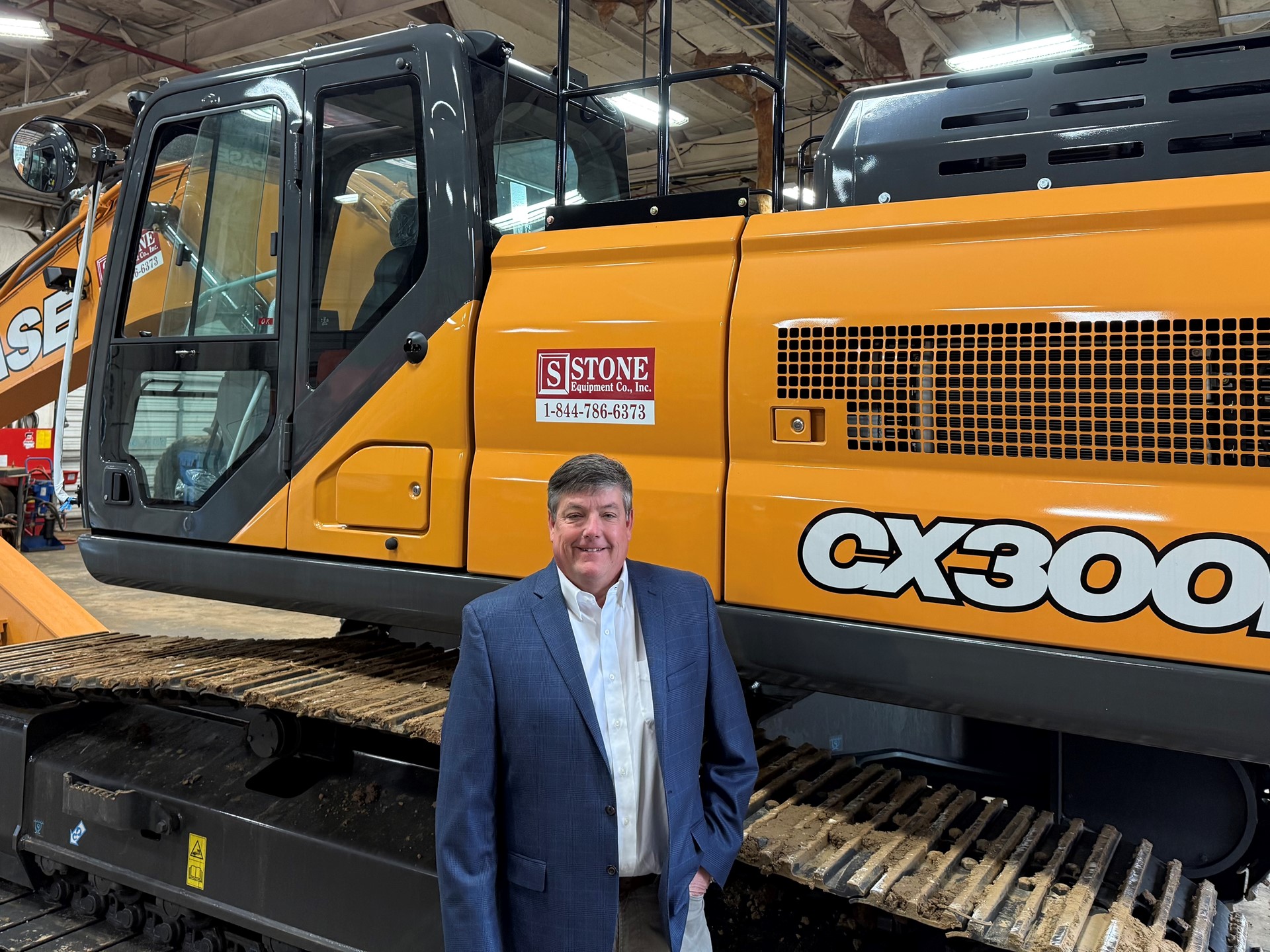 A man in a suit stands in front of a large orange Case CX300E excavator inside a warehouse. The excavator features a prominent logo and is positioned on a track, showcasing its size and design.
