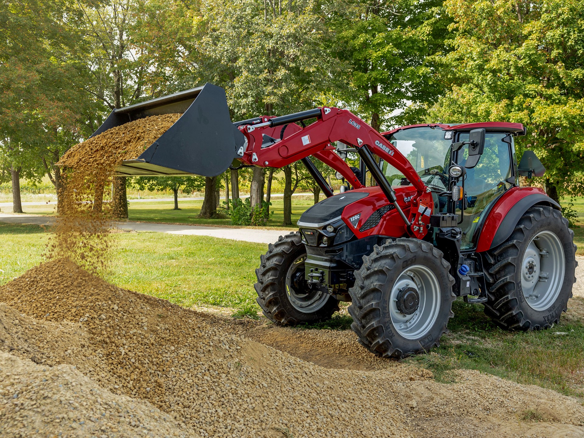 A red tractor with a front loader is dumping a pile of gravel onto the ground in a green outdoor setting. The tractor is positioned on a dirt path surrounded by trees, showcasing a sunny day. The loader is raised, and gravel is cascading down from the bucket.