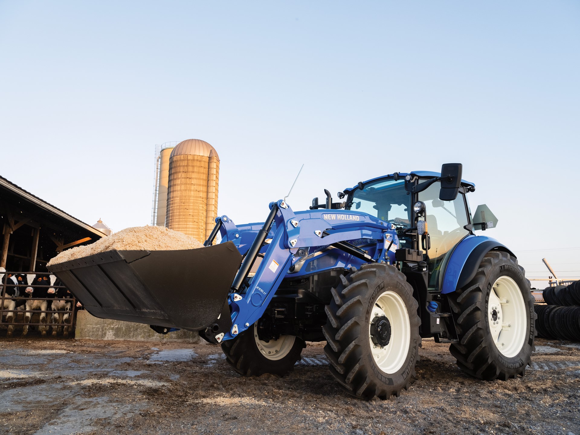 A blue New Holland tractor with a front loader is positioned on a farm, carrying a large bucket of material. In the background, a silos and a barn are visible under a clear sky, indicating a rural agricultural setting. The tractor's tires are large and rugged, suitable for farm work.