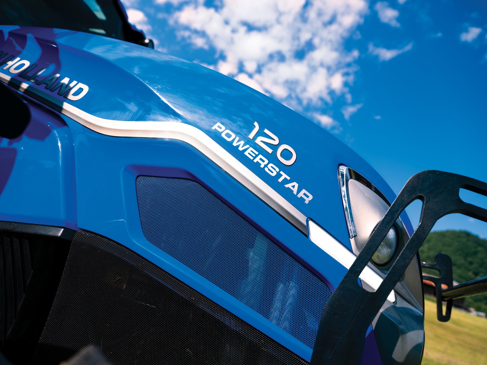 Close-up view of a blue New Holland tractor with the model name "120 Powerstar" prominently displayed. The image captures the tractor's sleek design against a backdrop of a clear blue sky with scattered clouds, highlighting its modern features and agricultural utility.