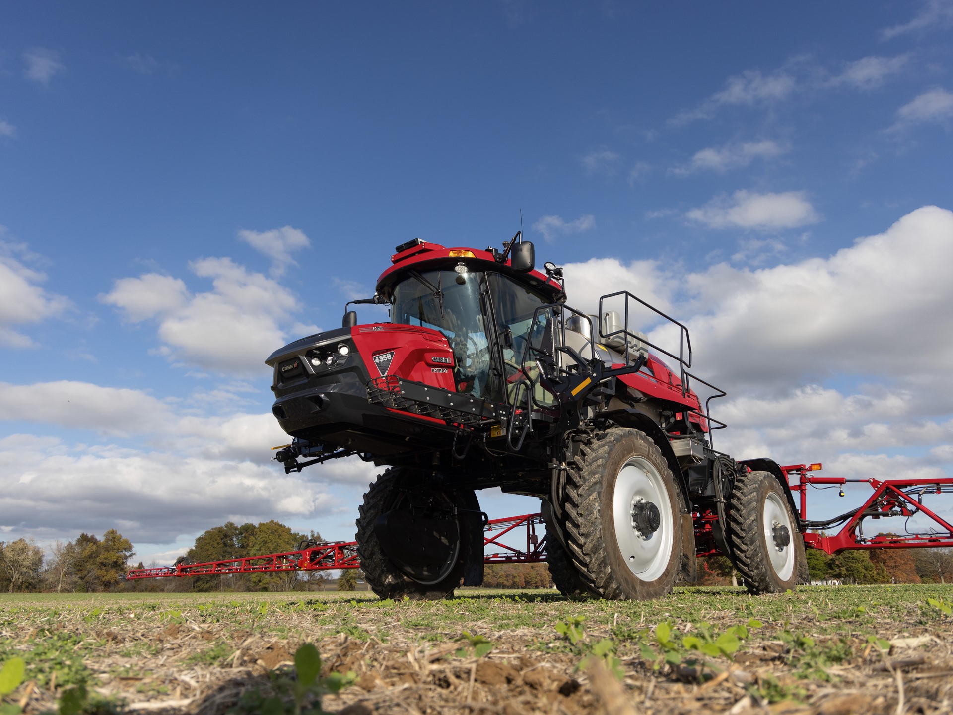 A red agricultural sprayer vehicle parked on a field under a partly cloudy sky. The sprayer features large wheels, a high cabin, and is equipped for crop spraying, showcasing its modern design and functionality.