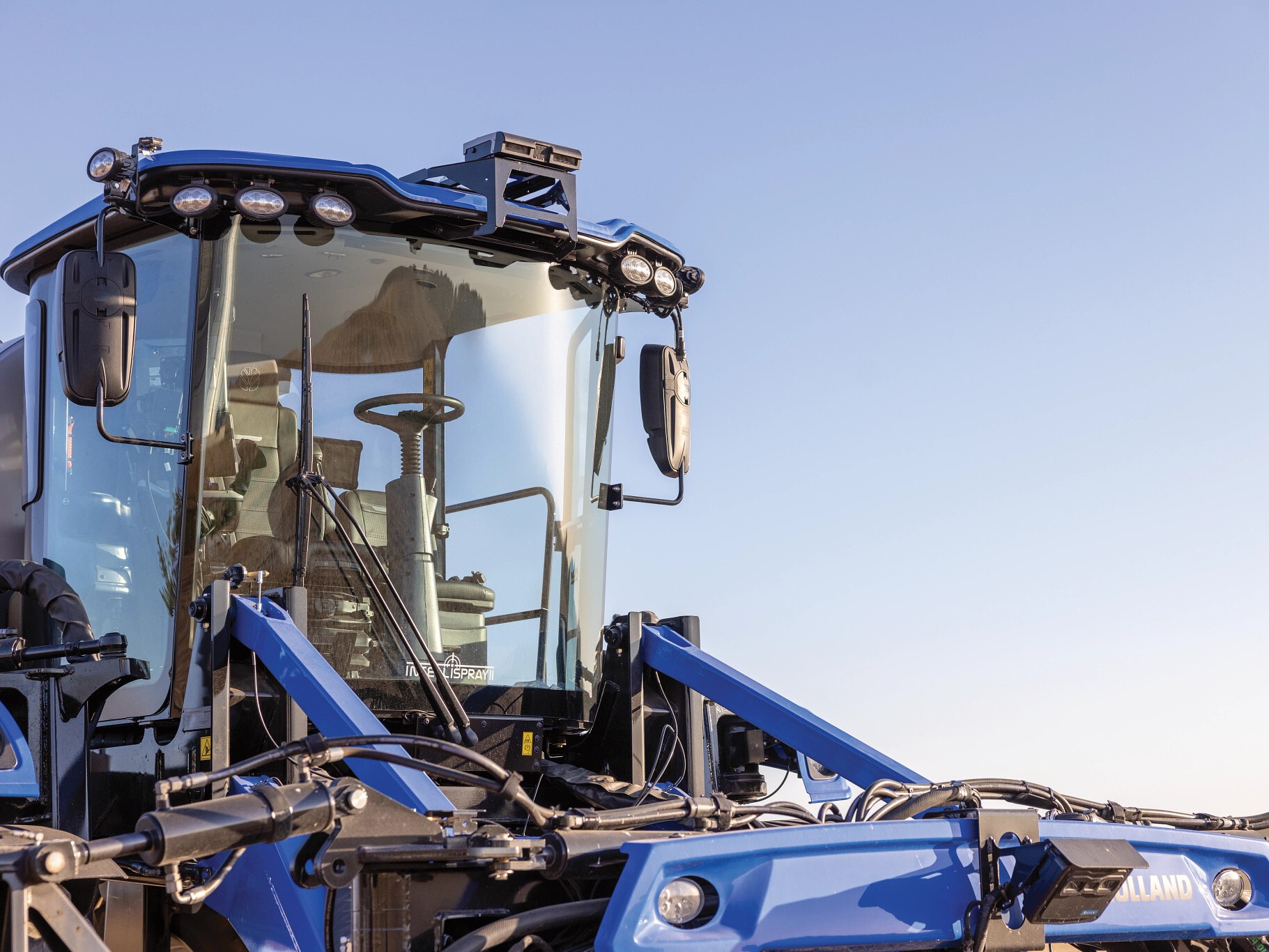 Close-up view of a modern blue agricultural tractor, showcasing its cab with large windows, advanced lighting, and various hydraulic components. The background features a clear blue sky, emphasizing the tractor's design and functionality for farming tasks.