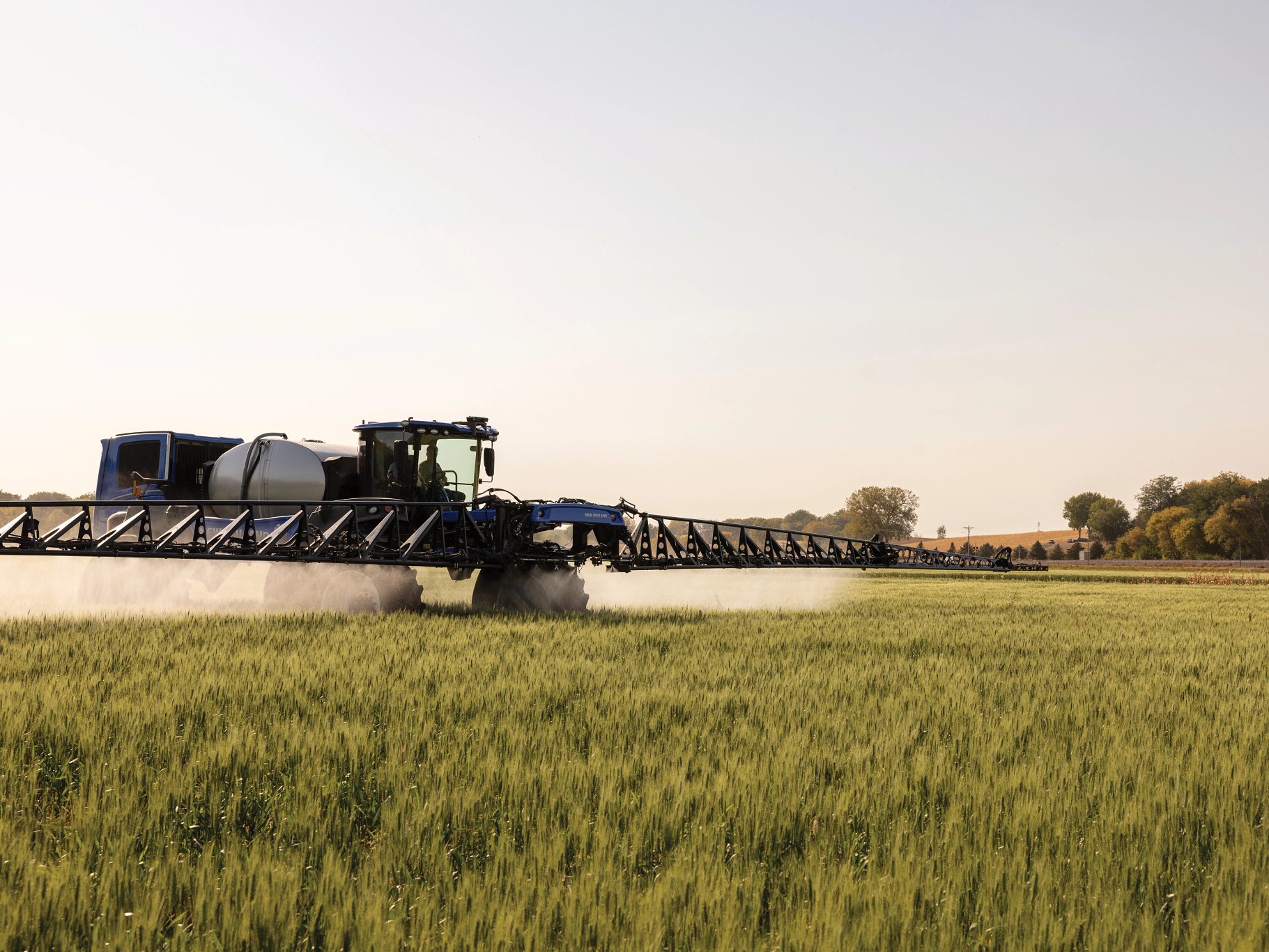 A blue agricultural tractor equipped with a large sprayer is working in a green field during a clear day. The tractor is spraying a mist over the crops, with trees and a clear sky in the background.