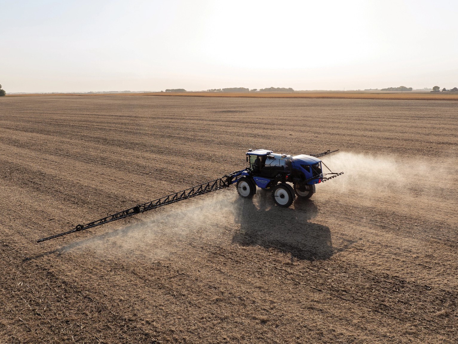 A blue tractor equipped with a sprayer is working in a vast, empty agricultural field. Dust is being kicked up as the tractor moves across the dry, brown soil under a clear sky. The landscape is flat and stretches into the distance, showcasing the agricultural setting.