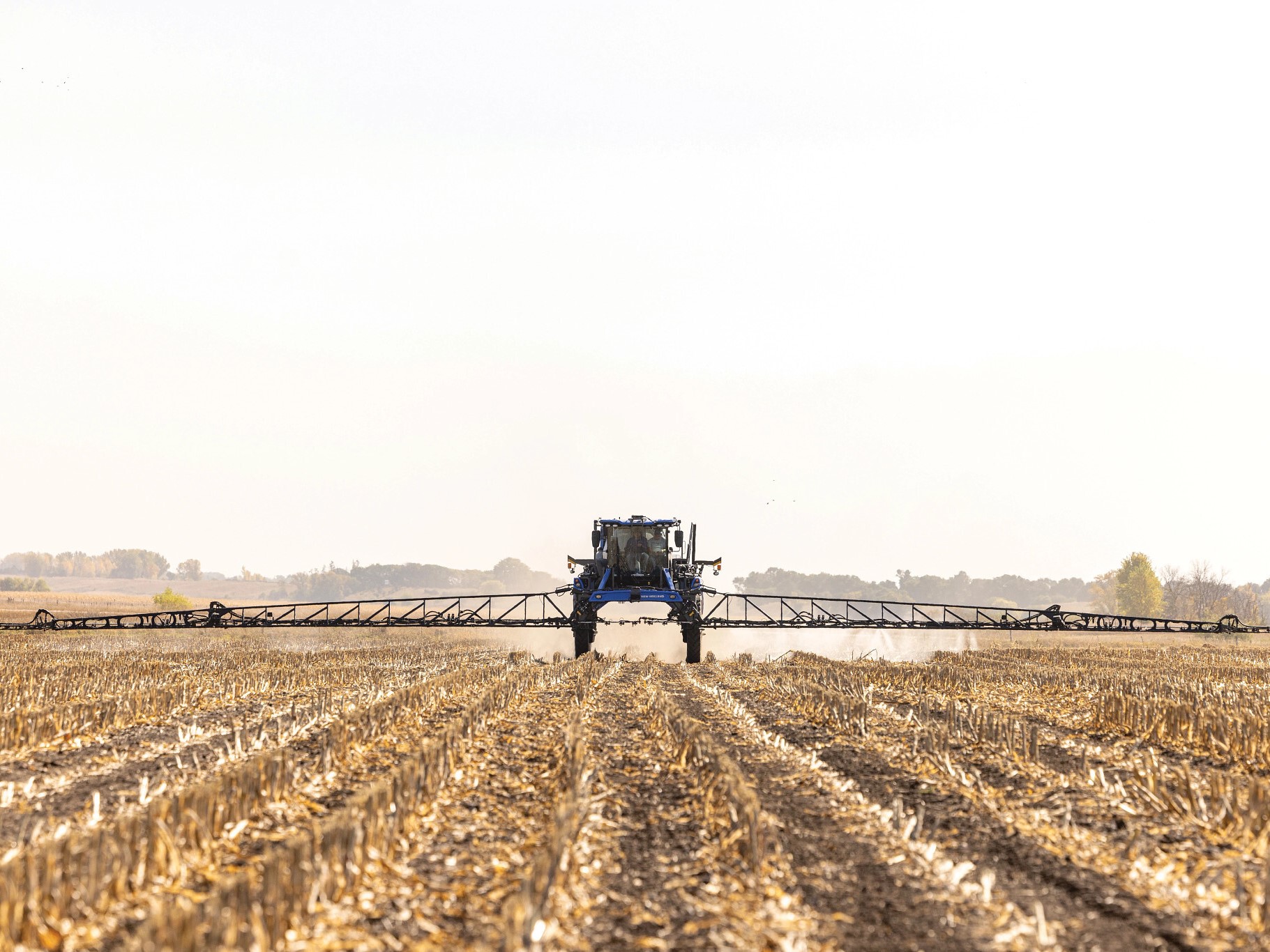 A large agricultural sprayer machine operates in a field, applying chemicals to the soil. The landscape features rows of harvested crops, with a clear sky in the background, indicating a sunny day.