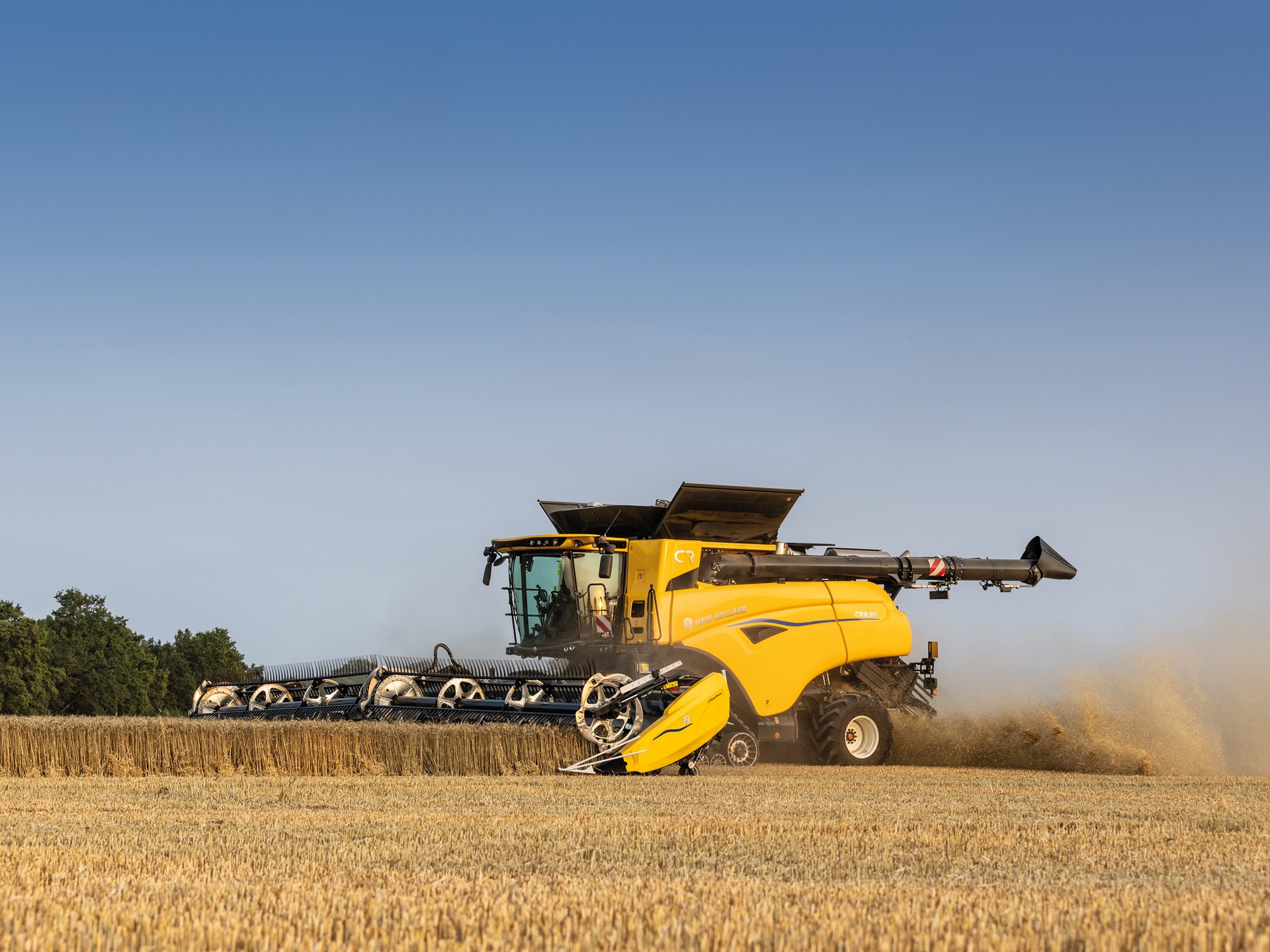 A yellow combine harvester working in a field during harvest season, with dust rising as it cuts through golden wheat under a clear blue sky.