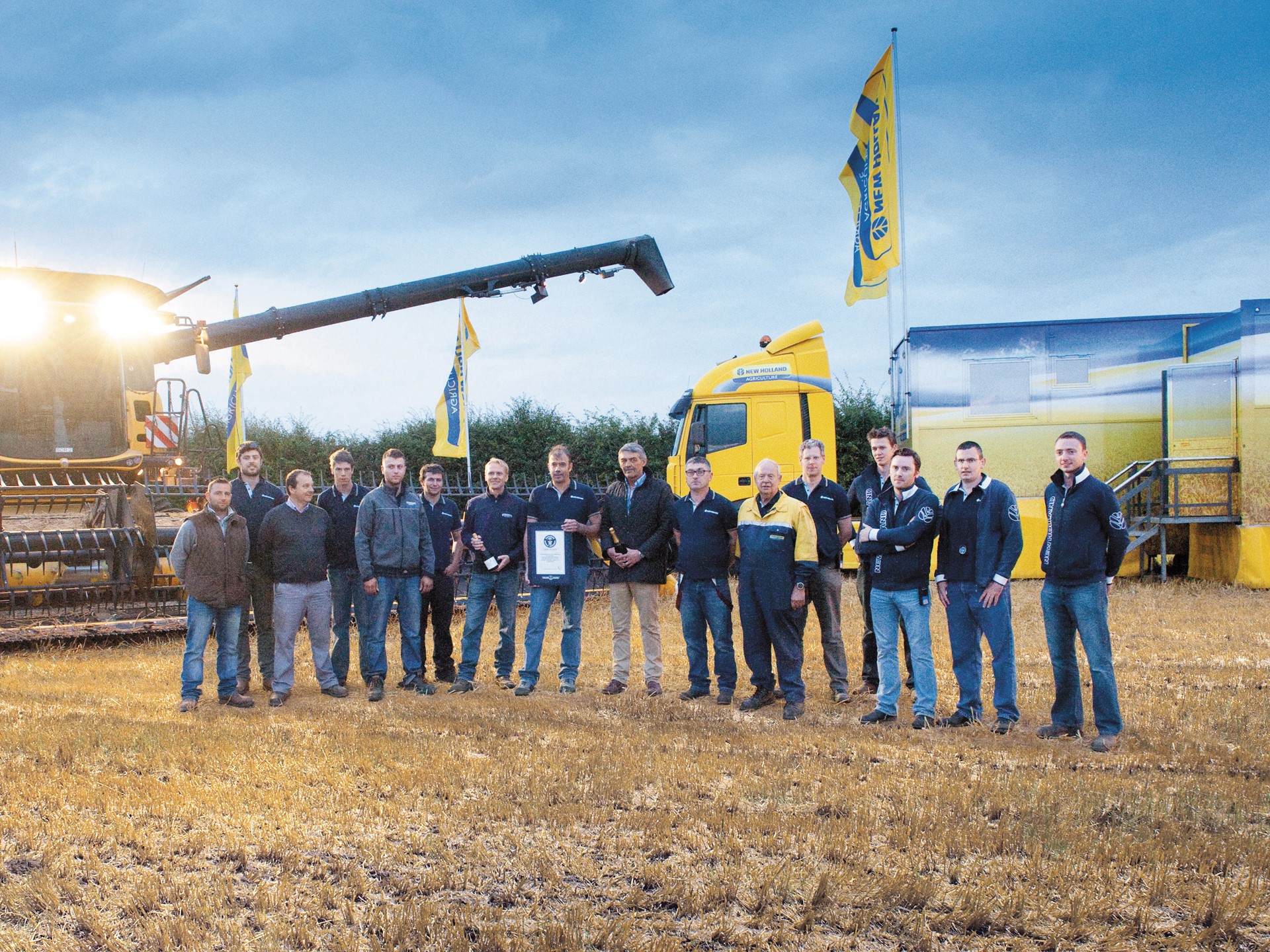 A group of 15 people stands in a field during twilight, with a large agricultural machine and a truck in the background. The individuals are dressed in casual work attire and are smiling, holding a plaque. Yellow flags with a logo are visible, indicating a celebration or event related to farming or agriculture. The setting appears to be a harvest or agricultural demonstration.