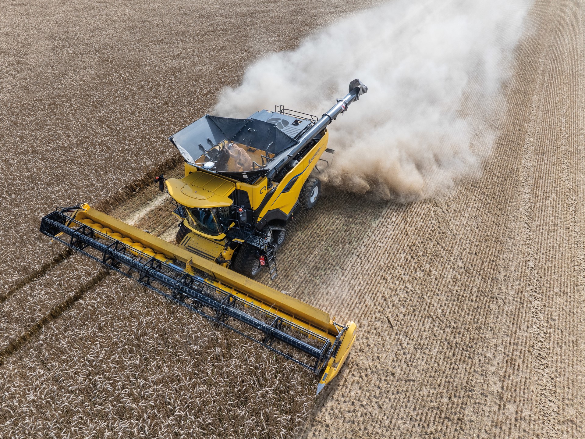Aerial view of a yellow combine harvester working in a field, with dust and debris being kicked up as it cuts through rows of crops. The machine is equipped with a wide header, efficiently harvesting the agricultural produce. The surrounding field shows neatly arranged crop rows, highlighting the agricultural landscape.