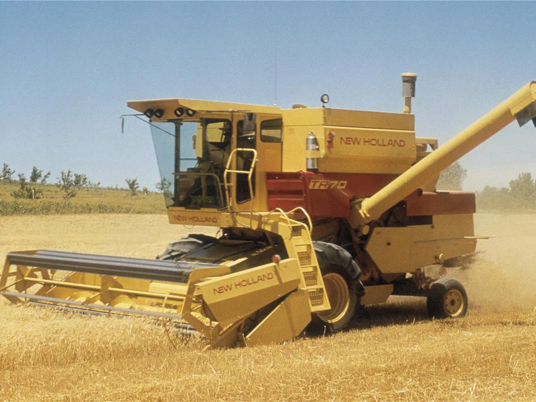 A New Holland TF70 combine harvester working in a golden wheat field, with dust rising as it cuts through the crops under a clear blue sky.