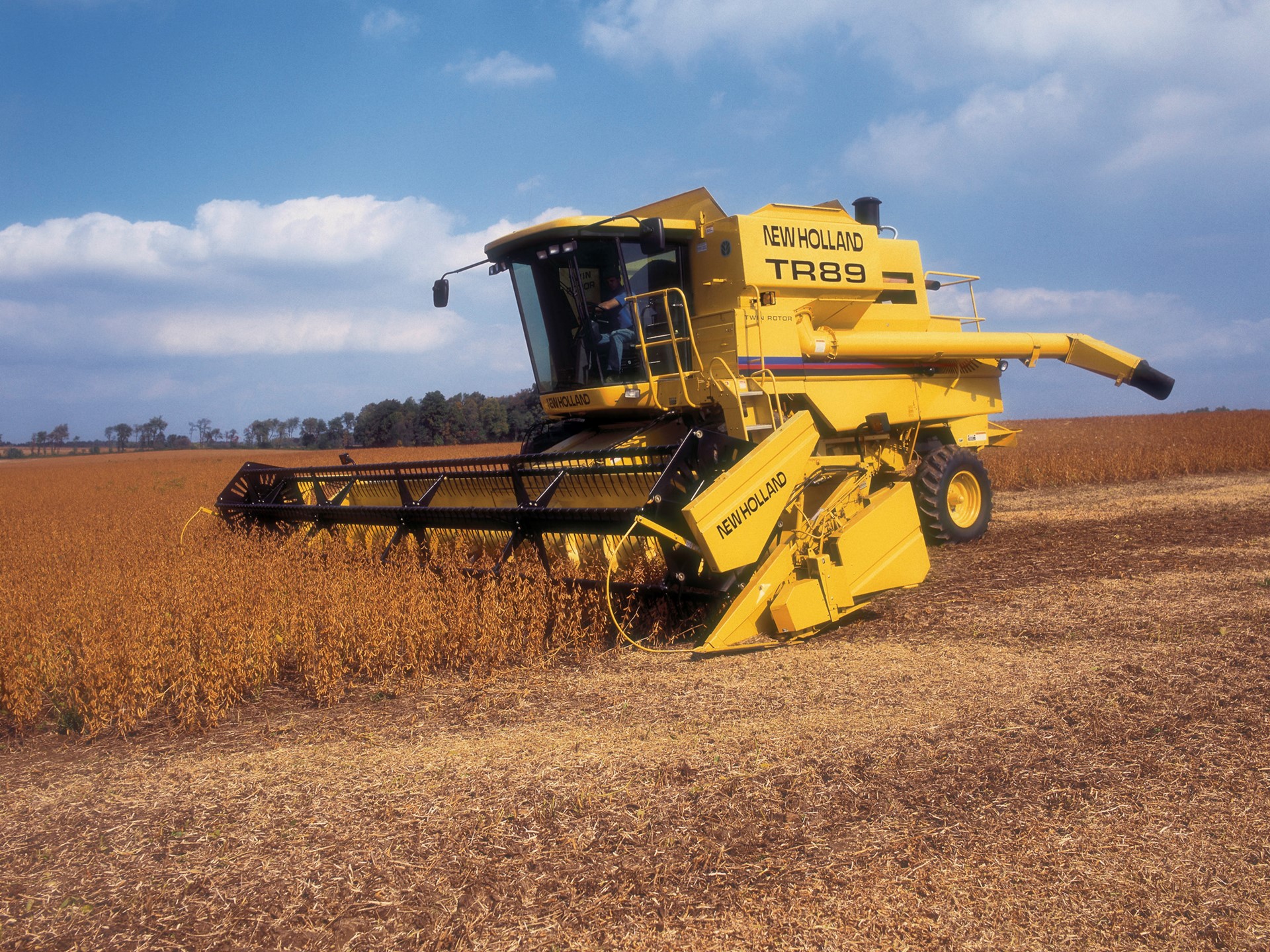 A yellow New Holland TR89 combine harvester working in a soybean field under a blue sky with scattered clouds. The machine is cutting through tall crops, showcasing agricultural machinery in action.