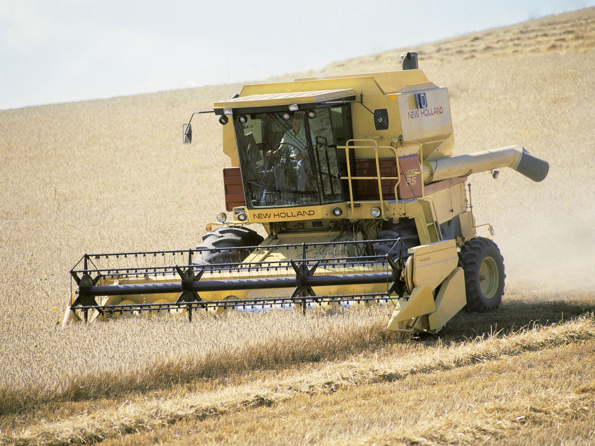 A yellow New Holland combine harvester working in a golden wheat field under a clear blue sky. The machine is cutting through the crops, with a farmer visible inside the cab, showcasing agricultural machinery in action.