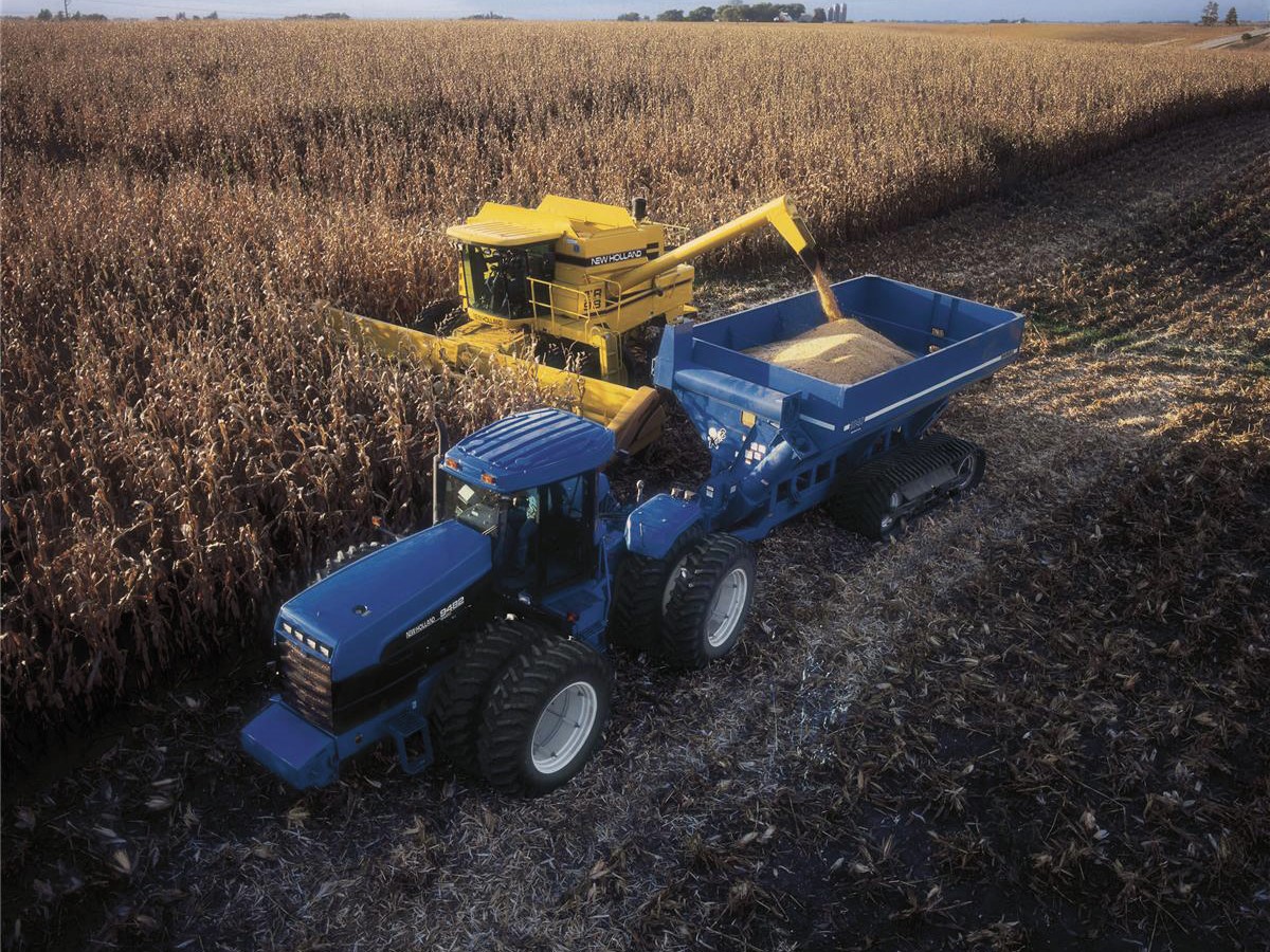 A blue tractor and a yellow combine harvester are working in a cornfield. The tractor is equipped with a large trailer, while the combine is harvesting corn. The landscape features rows of tall corn plants under a cloudy sky, showcasing an agricultural setting during the harvest season.