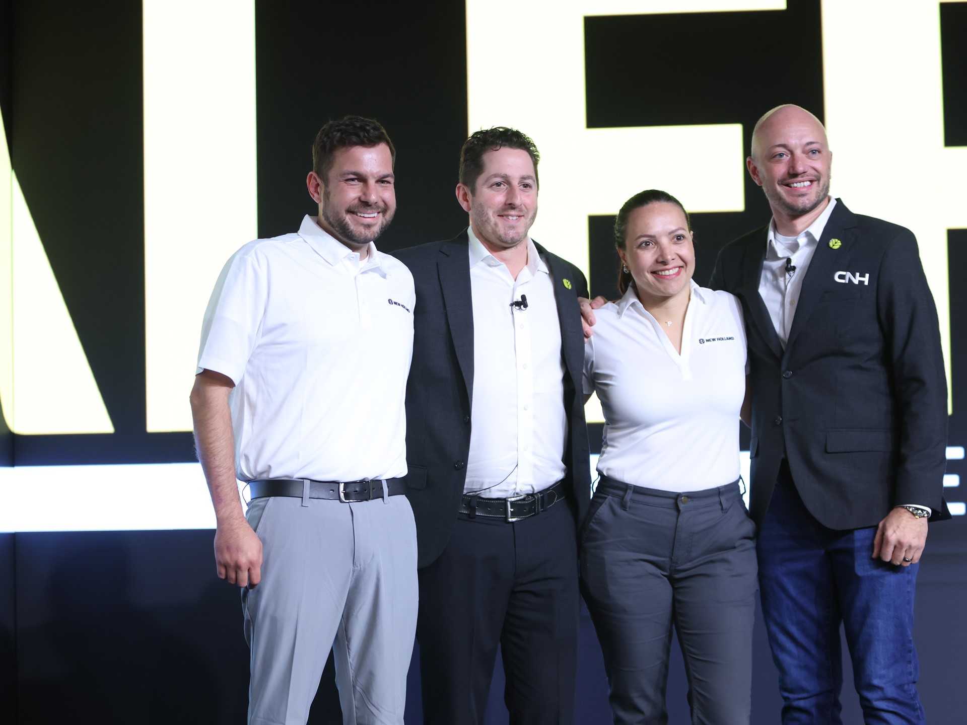 Four professionals pose together on stage at a corporate event with a large sign in the background They are dressed in business attire with two wearing white polo shirts and the others in a suit and a formal shirt The setting suggests a business meeting or conference