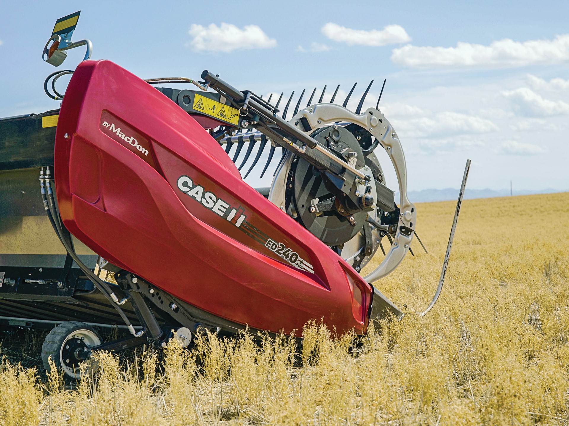 A close-up view of a red Case IH FD240 agricultural harvesting machine by MacDon operating in a golden crop field under a partly cloudy blue sky. The machine's cutting and gathering components are visible, with dry crops surrounding it.