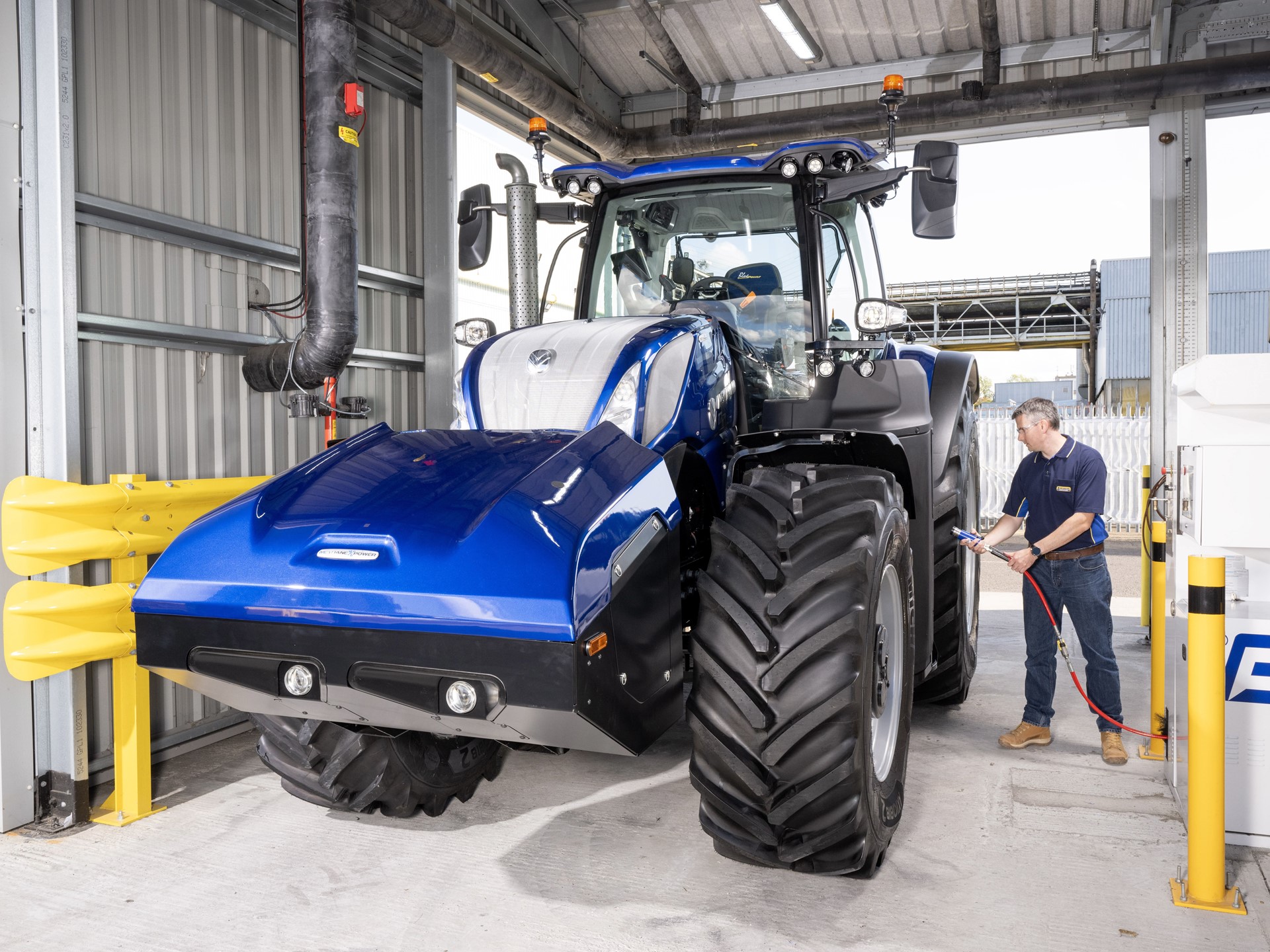 A man in a blue shirt and jeans is inspecting or charging a large blue agricultural tractor inside a modern industrial facility with metal walls and safety barriers.