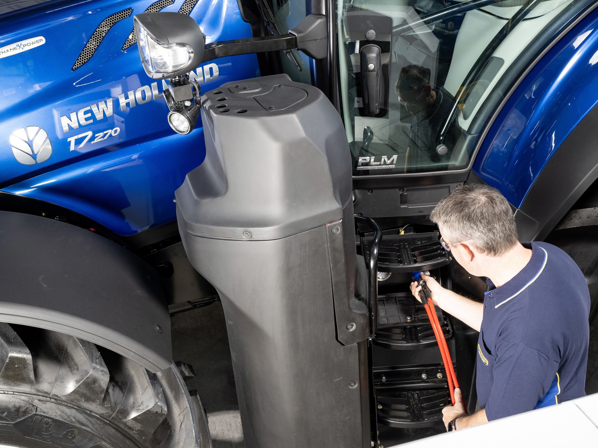 A man in a navy blue shirt is inspecting or repairing the steps of a blue New Holland T7.270 tractor inside a workshop or garage. The tractor's large front tire and side mirror are visible, along with the "Methane Power" label on the tractor's hood.