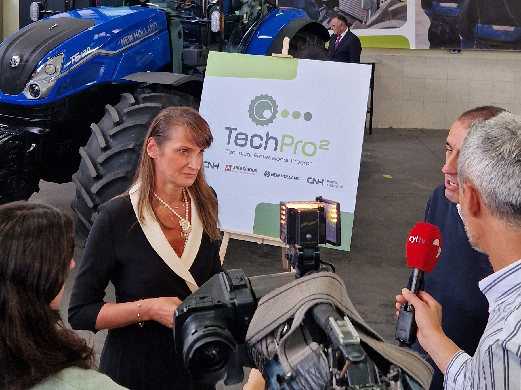 A woman is being interviewed by a news crew inside a workshop or showroom with a blue New Holland tractor in the background. Behind her is a large sign displaying the "TechPro2" logo and the text "Technical Professional Program." The setting includes posters of people working on machinery, emphasizing a technical or agricultural theme. The interviewers hold microphones and a professional video camera is recording the scene.