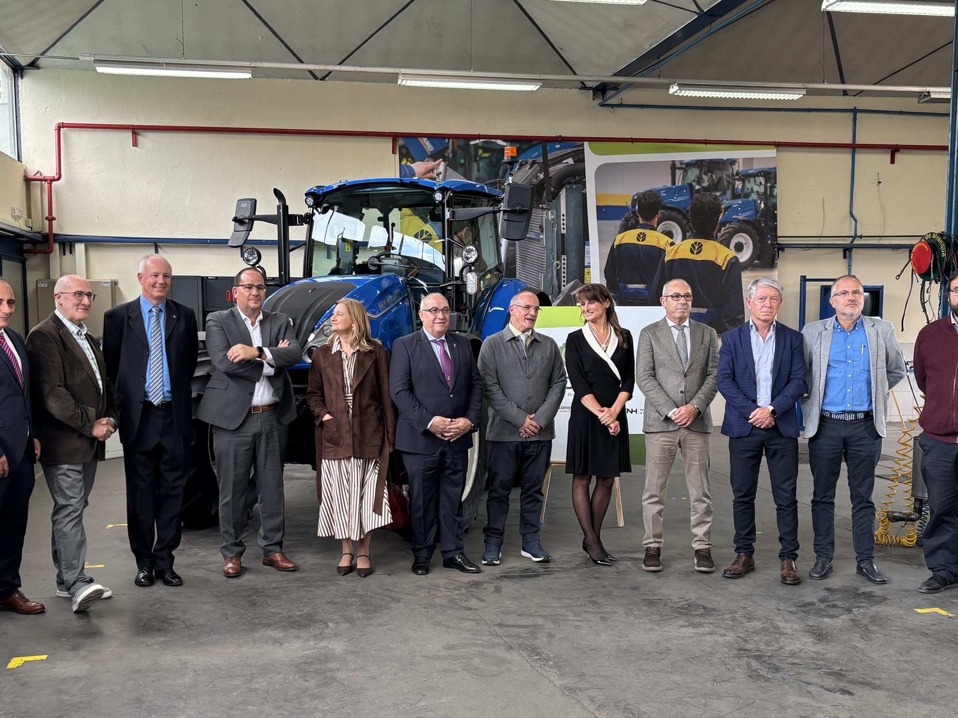 A group of twelve professionally dressed people standing in a row inside an industrial workshop with a large blue tractor behind them. The setting appears to be a formal event or visit, with a mix of men and women posing for a group photo. The background includes industrial equipment, posters, and workshop lighting.
