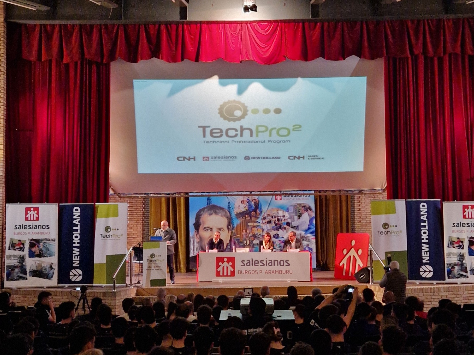 A large audience seated in an auditorium facing a stage with red curtains. On the stage, a speaker stands at a podium with a microphone, presenting in front of a large screen displaying the "TechPro² Technical Training Program" logo. Banners for Salesianos, New Holland, and CNH Industrial flank the stage. The event appears to be a formal presentation or training session.