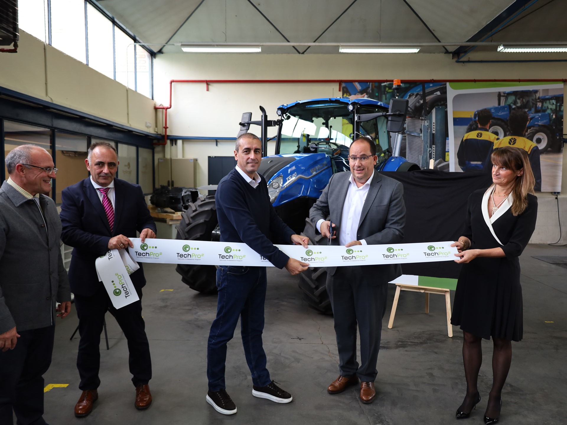 Five people standing in a workshop with a blue tractor in the background, participating in a ribbon-cutting ceremony. One man in a gray suit is cutting a white ribbon with "TechPro" logos, while the others hold the ribbon and smile. The setting appears to be an industrial or agricultural equipment facility.