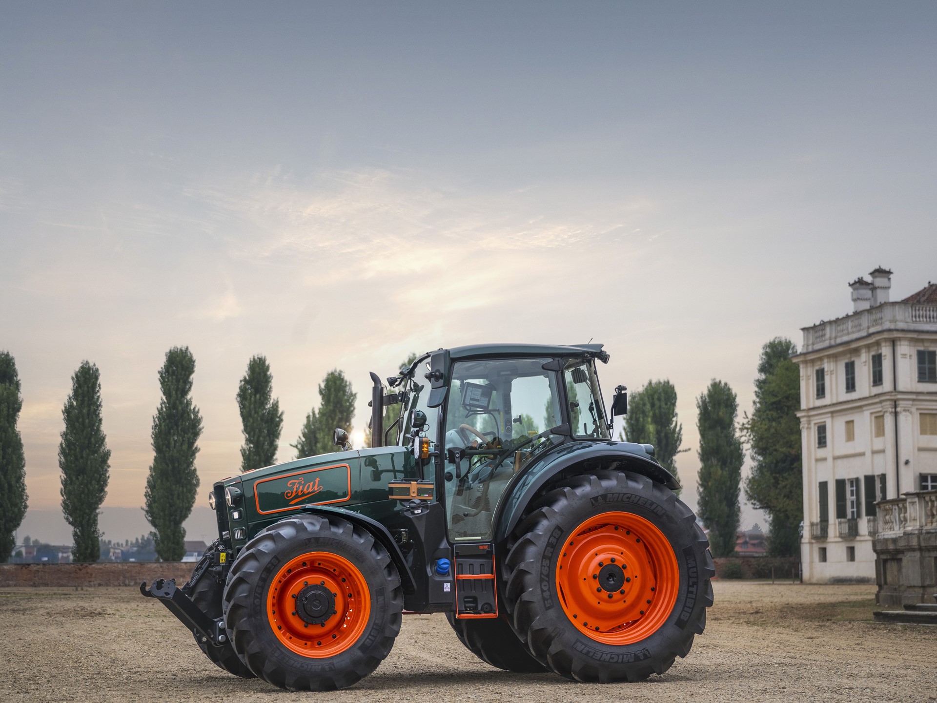 A green and orange Fiat tractor parked on a dirt ground with tall trees in the background and a large white building on the right side under a cloudy sky during sunset.