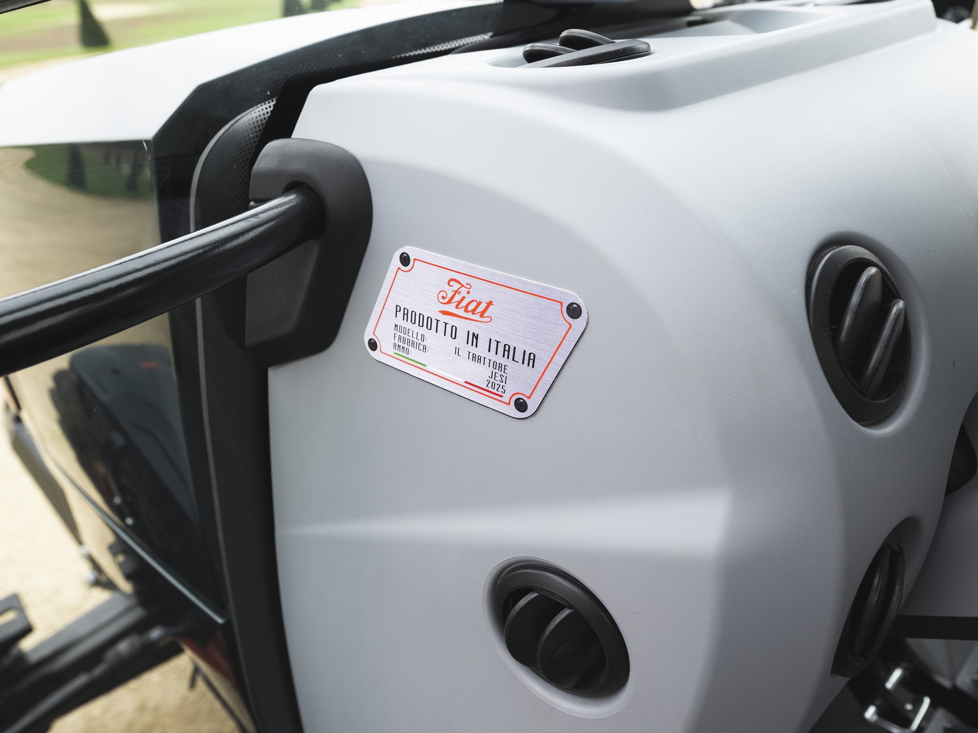 Close-up view of a gray Fiat tractor dashboard showing air vents and a metal label with the text "Fiat Prodotto in Italia" indicating the model and manufacturing details.
