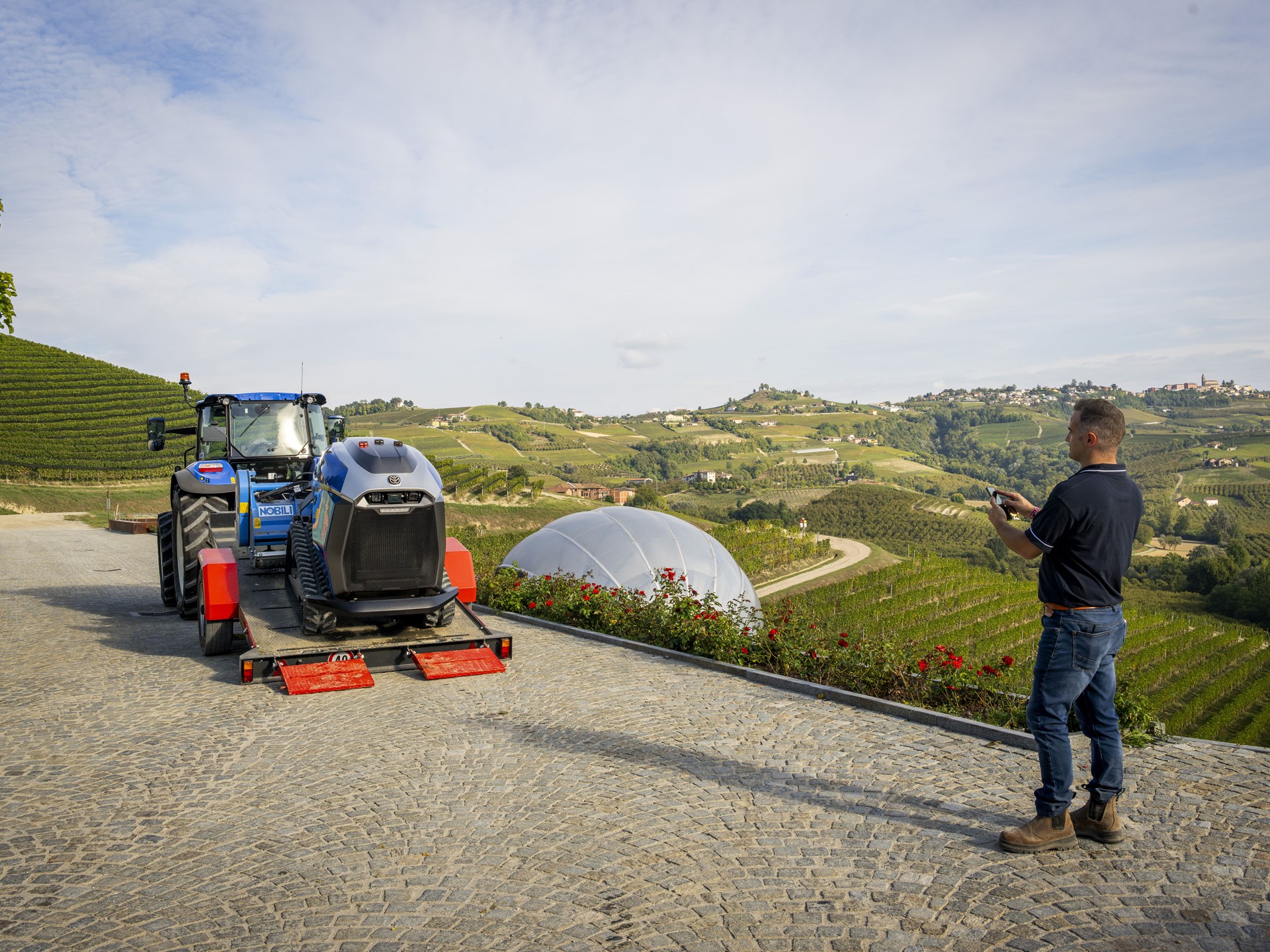 A man operating a remote control stands on a cobblestone path overlooking a scenic vineyard landscape with rolling hills. In front of him, a blue tractor is carrying a modern robotic agricultural machine on a trailer. The vineyard rows stretch across the hills under a partly cloudy sky, with a white dome structure visible among the greenery.