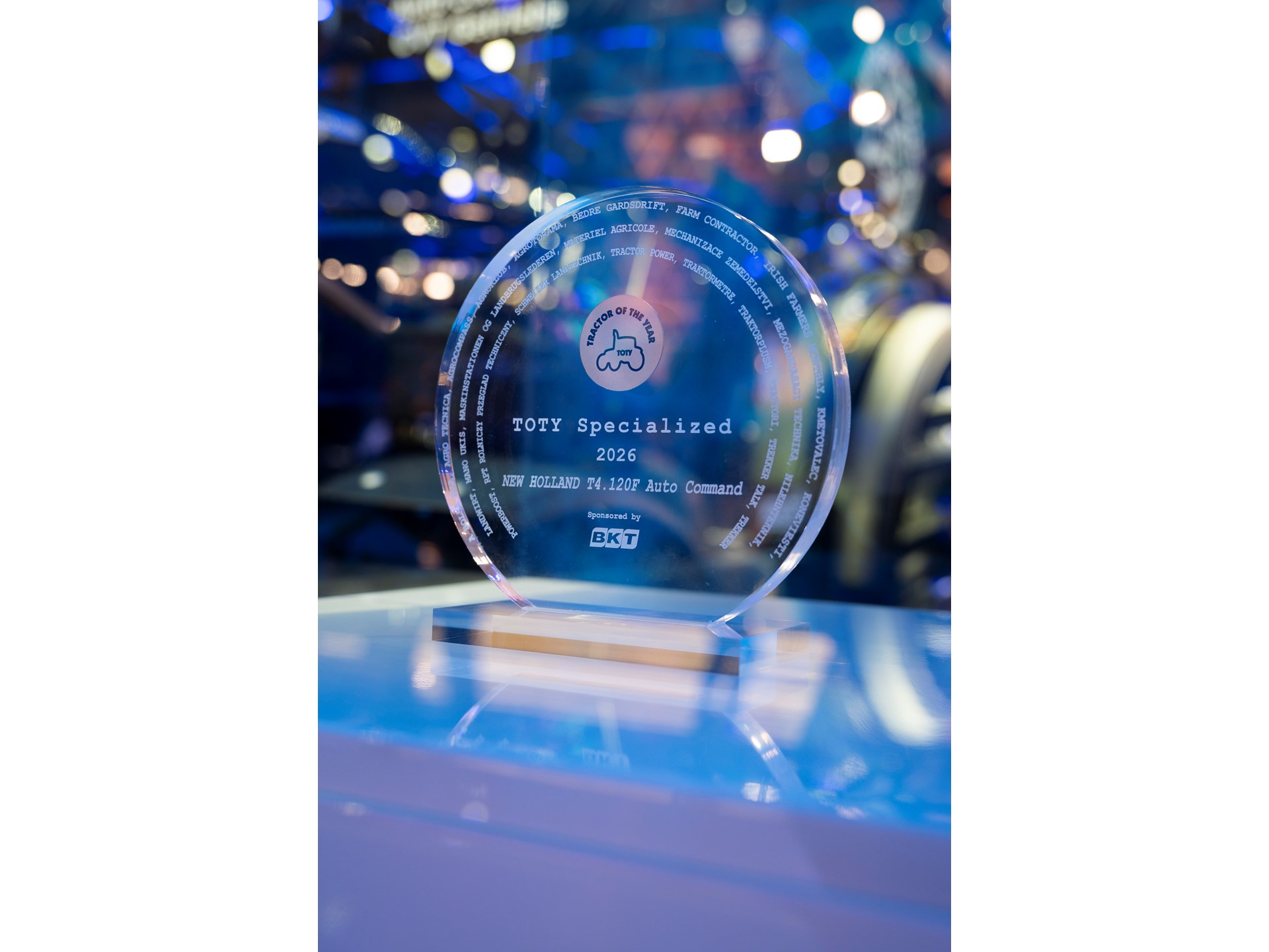 A clear circular glass award trophy with the inscription "TOTY Specialized 2026 NEW HOLLAND T4.120F Auto Command" and the logo "Tractor of the Year" featuring a tractor icon. The award is sponsored by BKT and is displayed on a reflective surface with a blurred background of blue and purple lights.