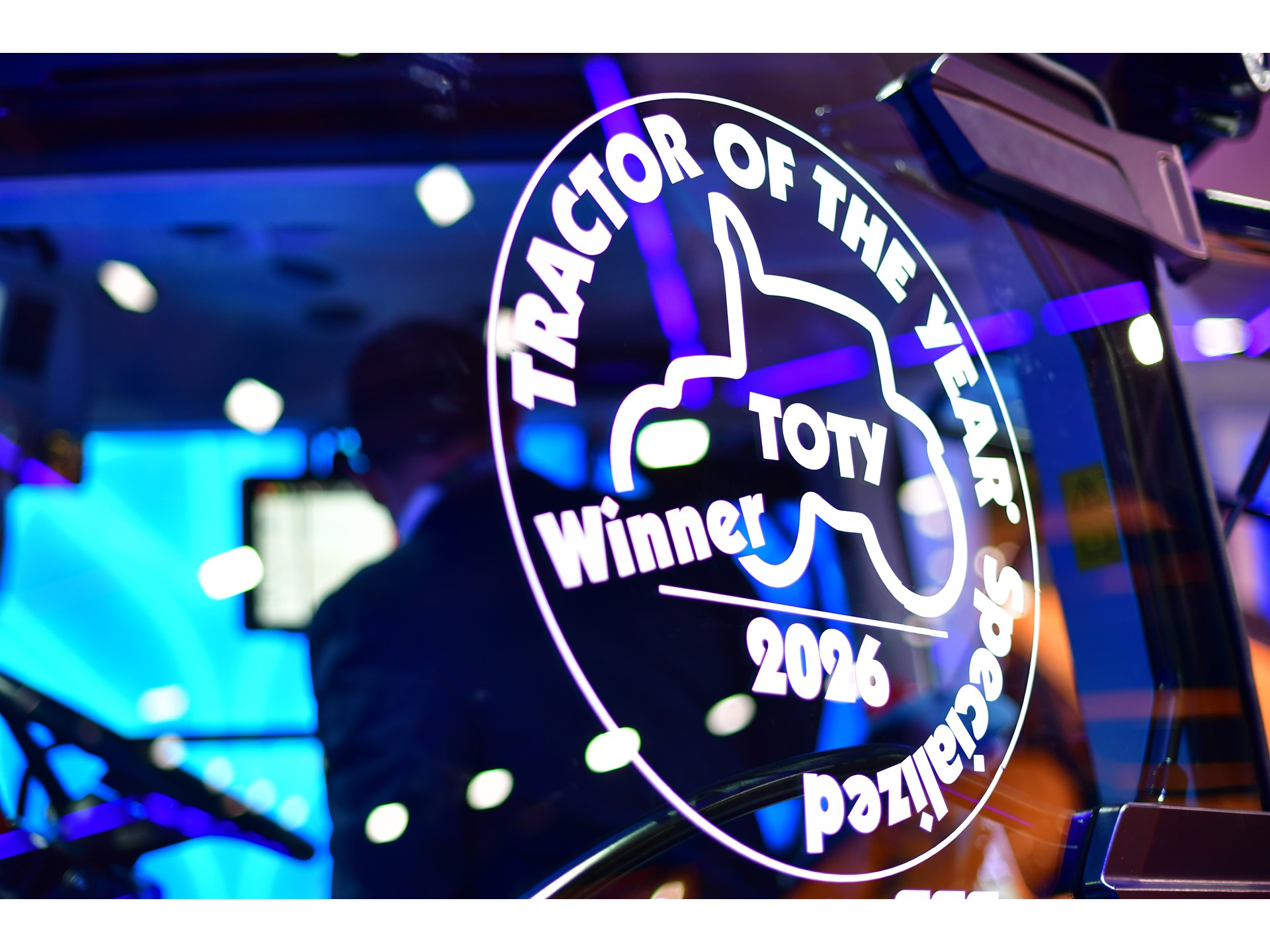 Close-up of a glass window on a tractor displaying a white decal that reads "TRACTOR OF THE YEAR Specialized Winner 2026 TOTY" with a tractor silhouette in the center, reflecting colorful blue and purple lights in the background.