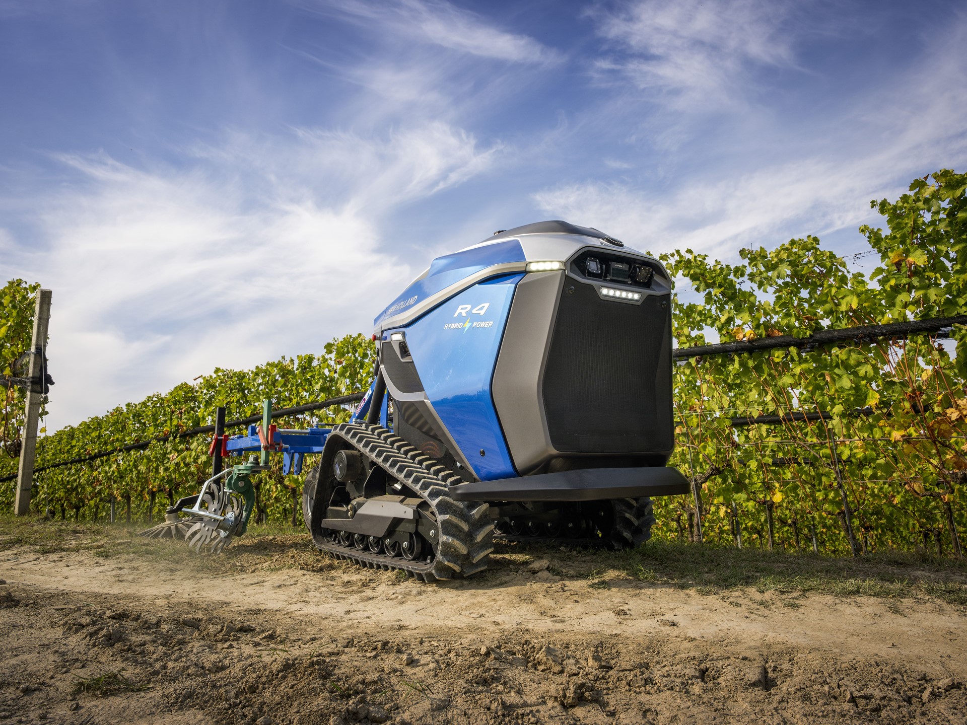 A blue autonomous agricultural robot with tank-like tracks is working on a dirt path next to rows of green grapevines under a partly cloudy sky. The robot is equipped with farming tools and appears to be designed for vineyard maintenance.