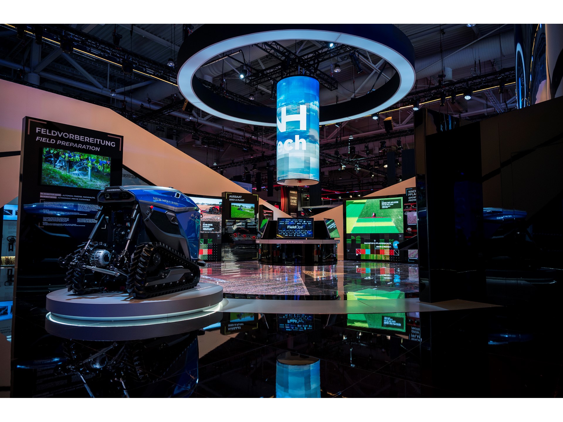 Exhibition booth showcasing advanced agricultural technology with a blue autonomous robotic vehicle on a circular platform, multiple digital screens displaying field preparation and seed planting information, and a large cylindrical hanging digital display with the word "Tech" in a modern indoor event space.