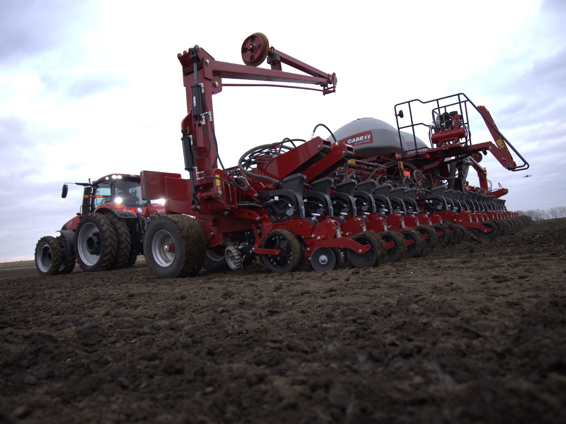 A large red agricultural tractor attached to a multi-row planter machine working on a freshly tilled field under a cloudy sky, preparing the soil for planting crops.