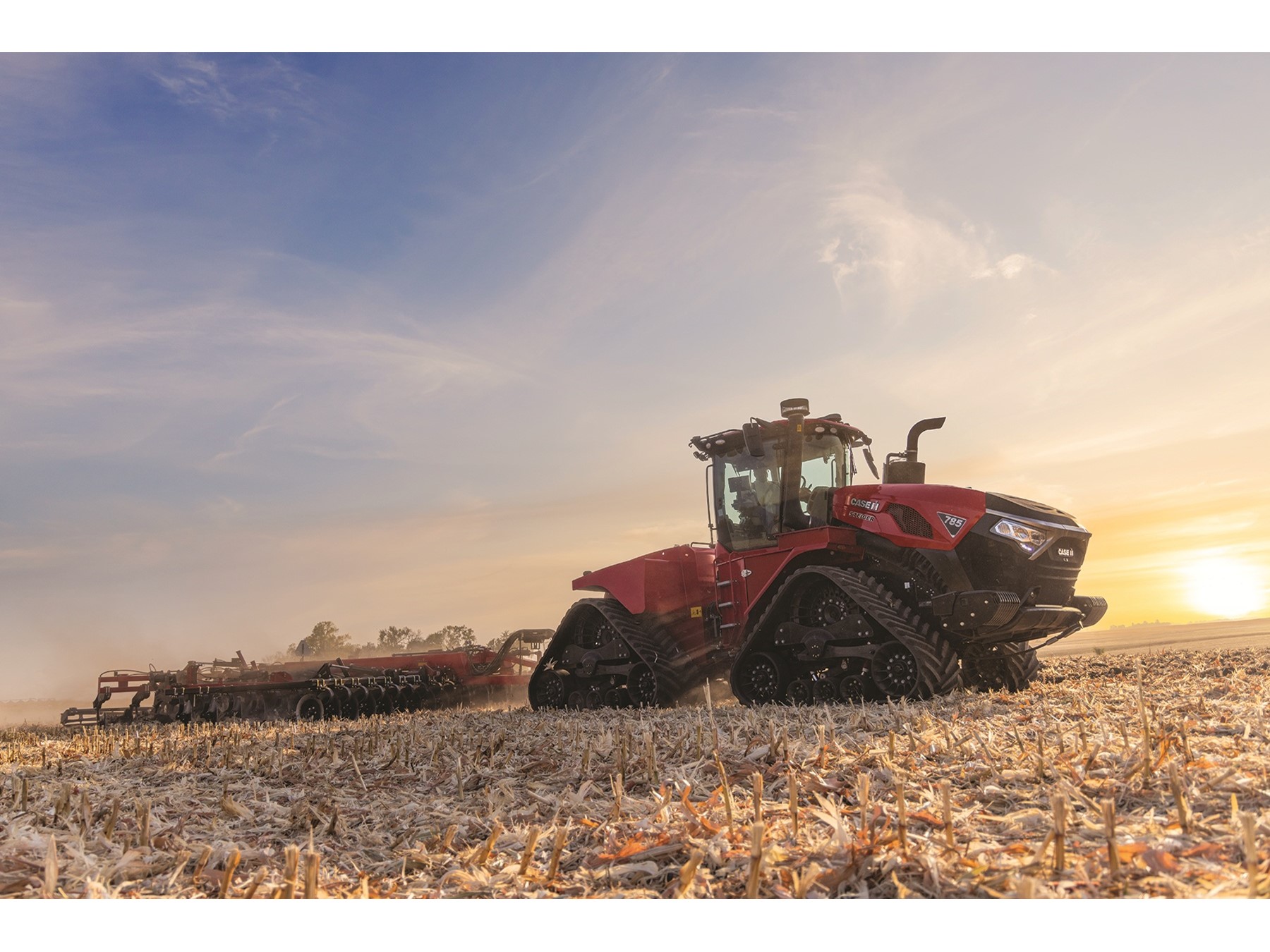 A large red Case IH tractor with rubber tracks is working in a harvested field during sunset, pulling farming equipment behind it under a partly cloudy sky.