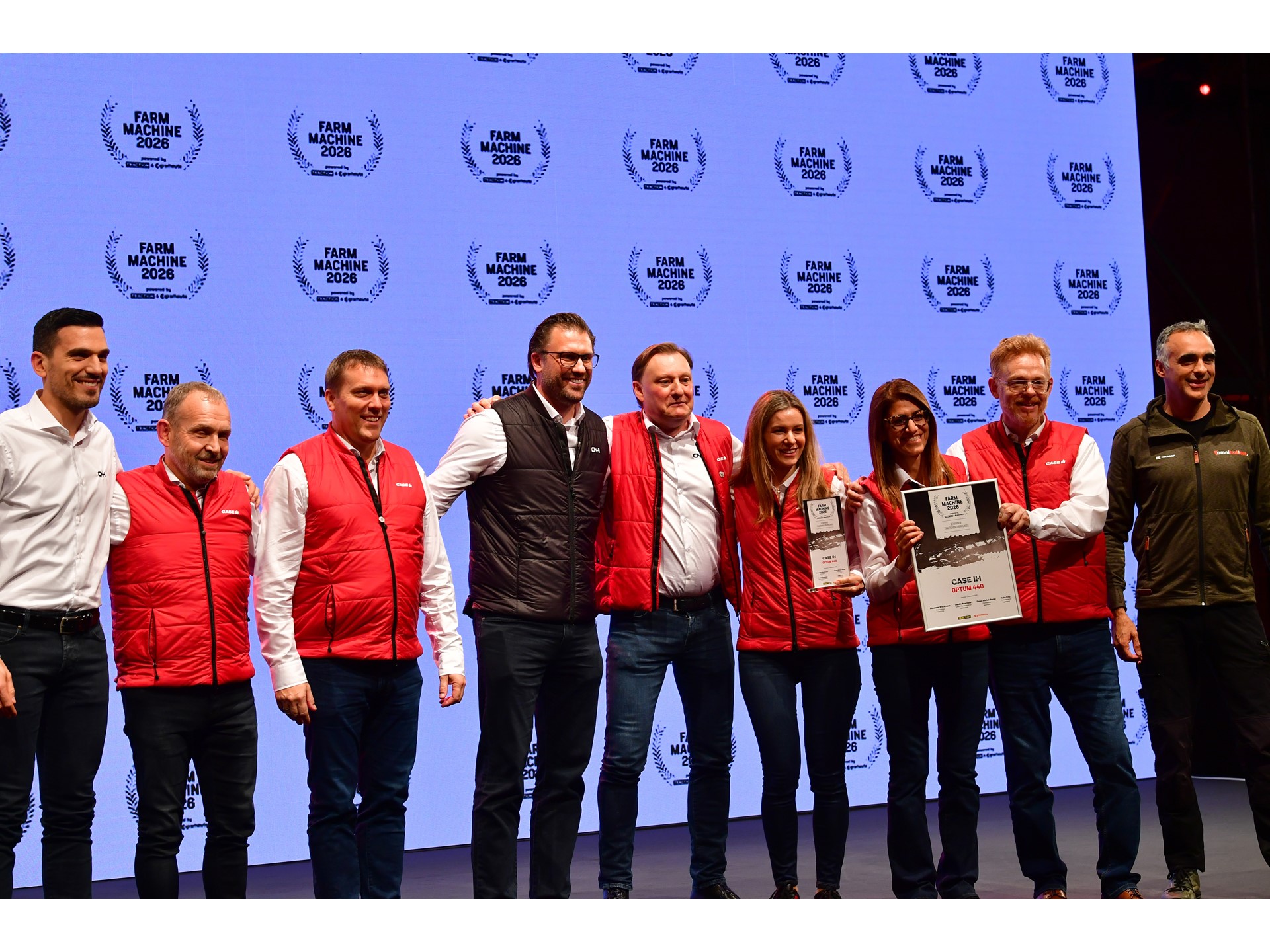 A group of nine people standing on stage in front of a large screen displaying the "Farm Machine 2026" logo repeatedly. Most individuals are wearing red vests over white shirts, while two are in different colored jackets. Two women in the center are holding awards, one holding a plaque and the other a certificate. The group is smiling and posing for a photo, suggesting a celebratory or award event.