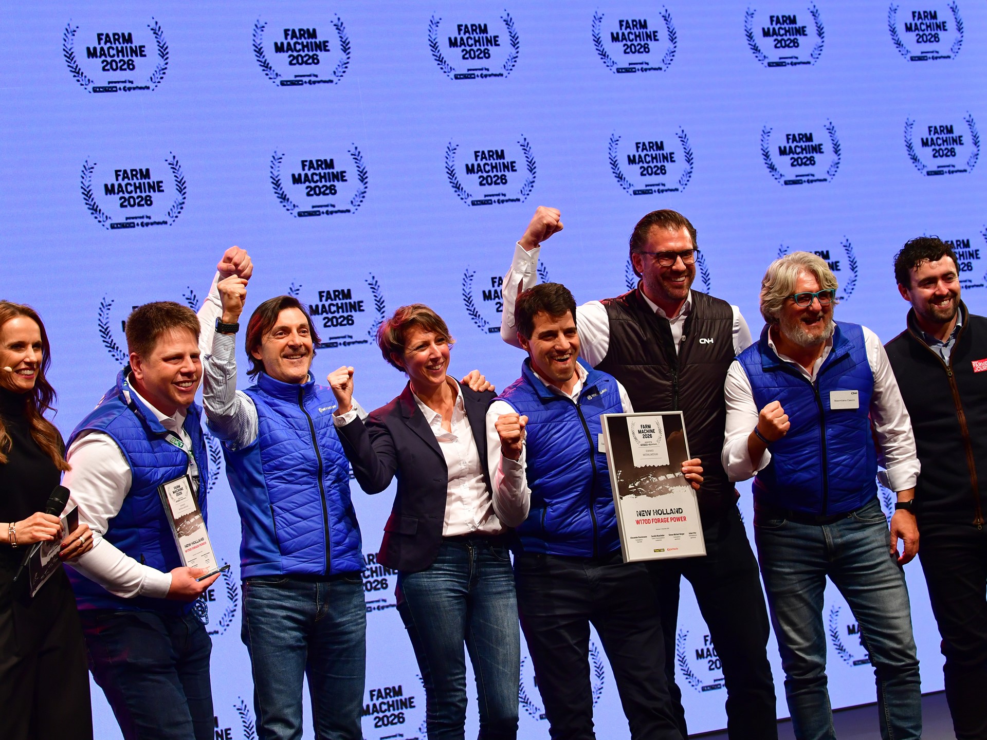 A group of eight people standing on stage celebrating an award at the Farm Machine 2026 event. Most of them are wearing blue vests and smiling, with some raising their fists in excitement. One person is holding a framed certificate that reads "New Holland Methane Power." The background features a repeated "Farm Machine 2026" logo.