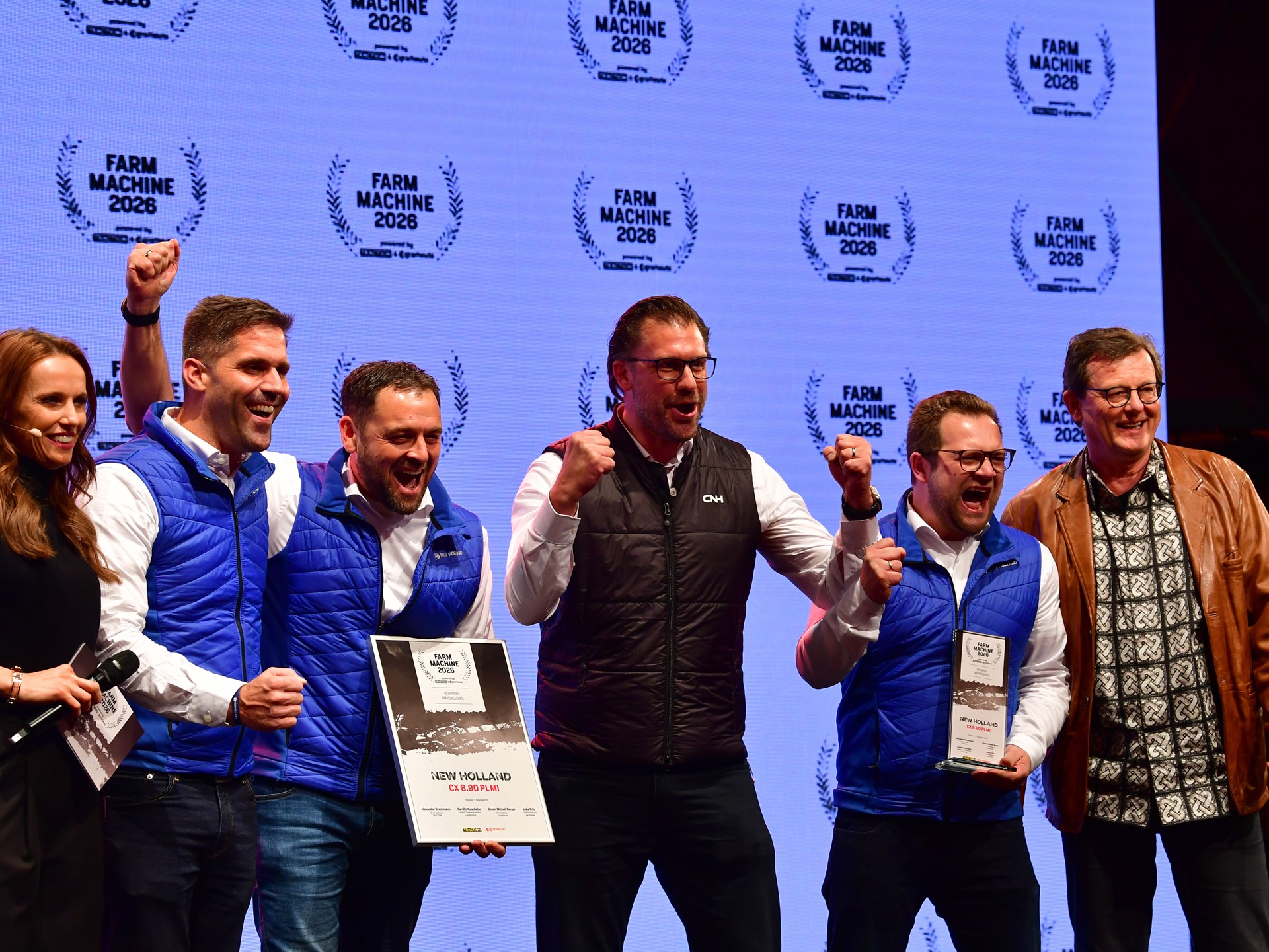 A group of six people standing on stage celebrating an award. Four men in blue and black vests are holding plaques and certificates, smiling and raising their fists in excitement. A woman on the left holds a microphone and a man on the right wears a brown leather jacket and patterned shirt. The background displays multiple "Farm Machine 2026" logos.
