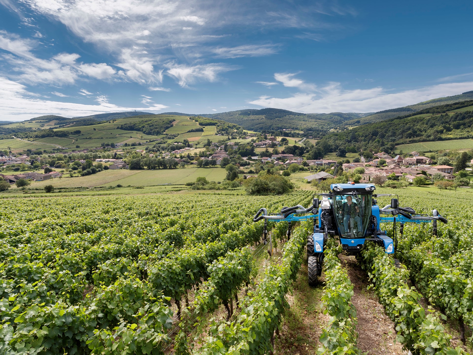 A blue agricultural tractor working in a lush green vineyard with rows of grapevines under a partly cloudy blue sky, with rolling hills and a small village in the background.