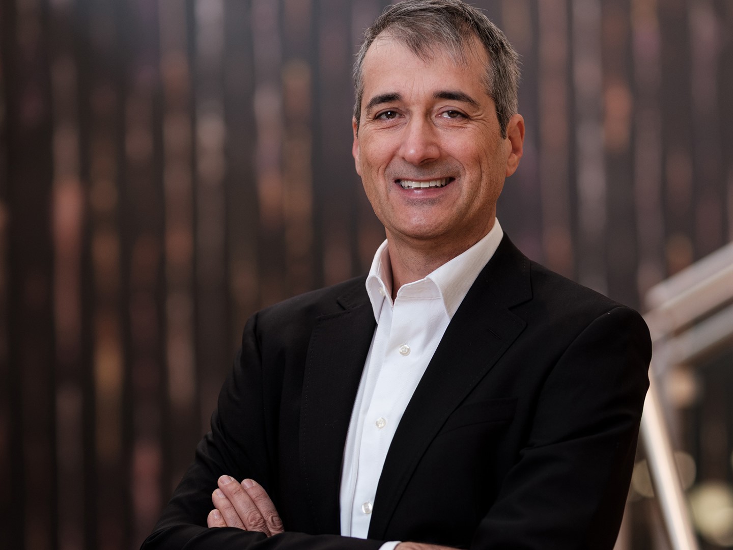 A middle-aged man with short gray hair wearing a black suit jacket and white dress shirt stands with his arms crossed, smiling confidently against a blurred dark brown and black background.
