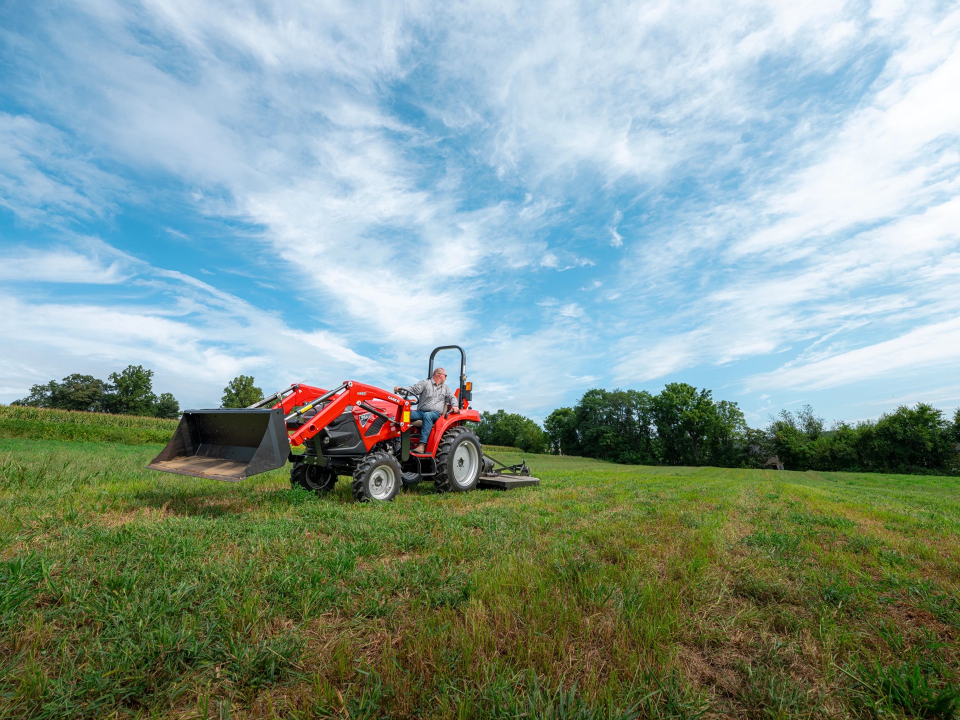A man driving a red tractor with a front loader attachment across a green grassy field under a partly cloudy blue sky, with trees in the background.