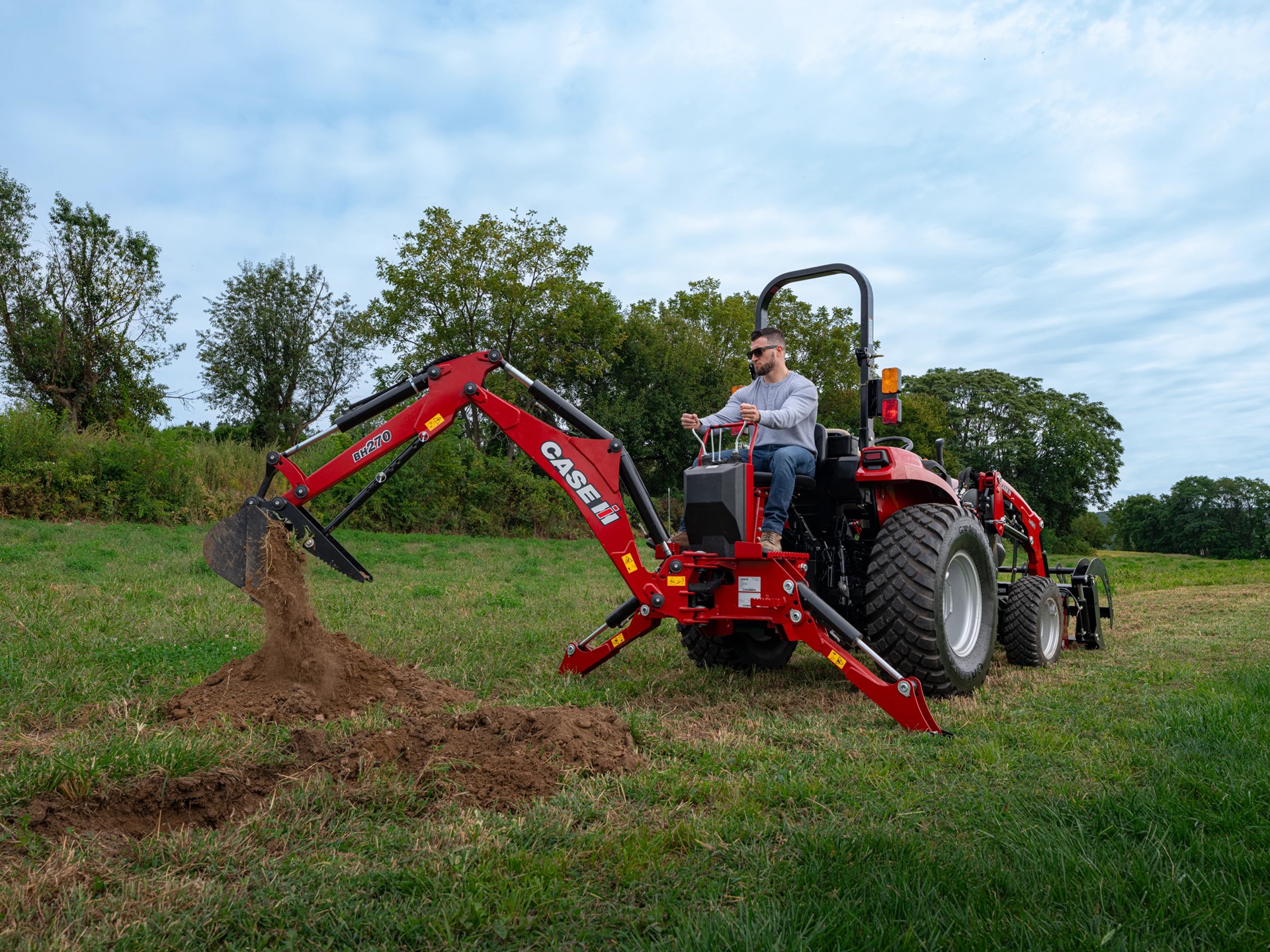 A man operating a red CASE IH tractor equipped with a backhoe attachment, digging into the grassy ground in a rural outdoor setting with trees and a cloudy sky in the background.