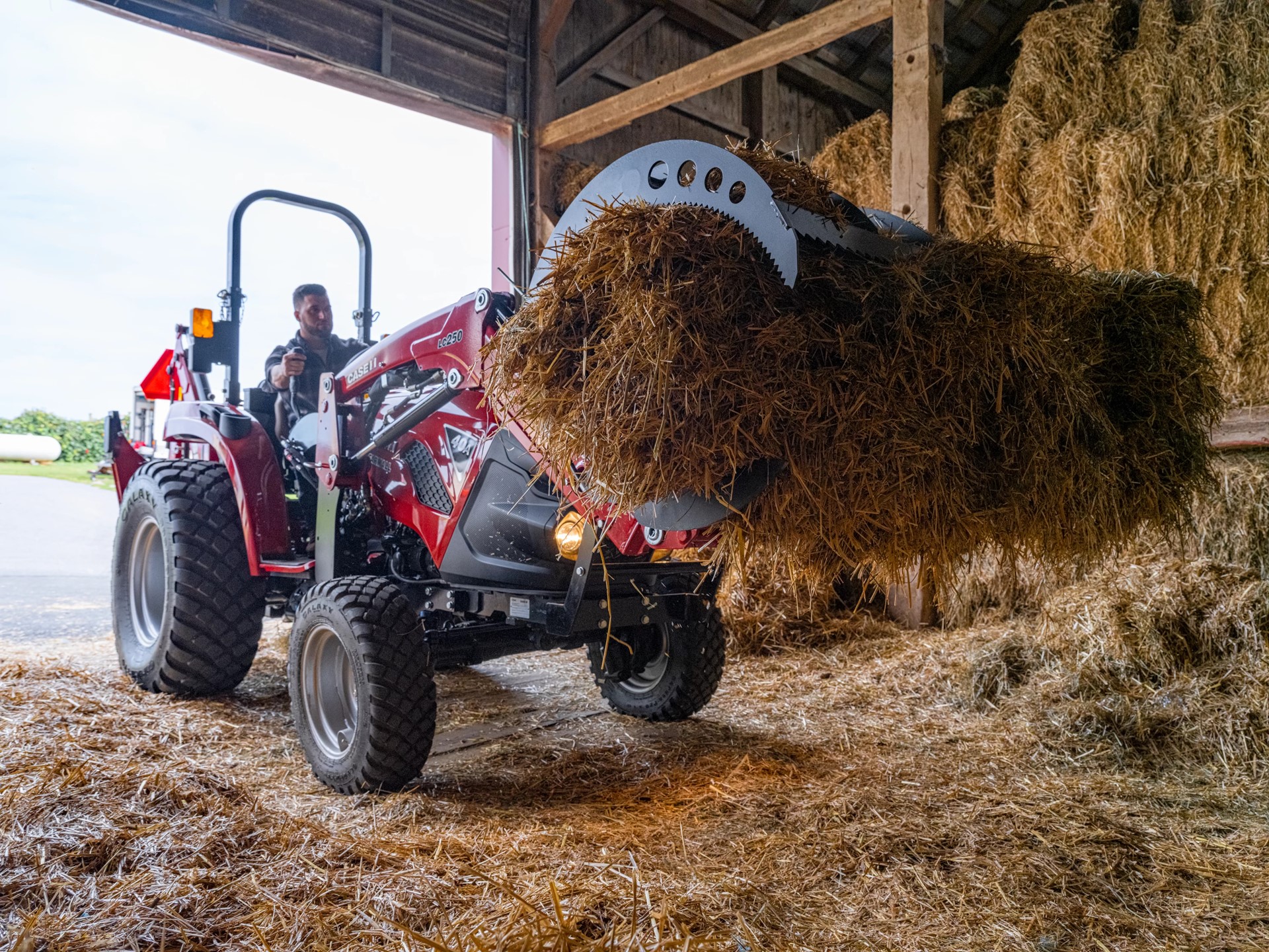 A person operating a red tractor inside a barn, using a front loader attachment to lift a large bale of hay. The barn is filled with stacked hay bales and straw scattered on the ground.