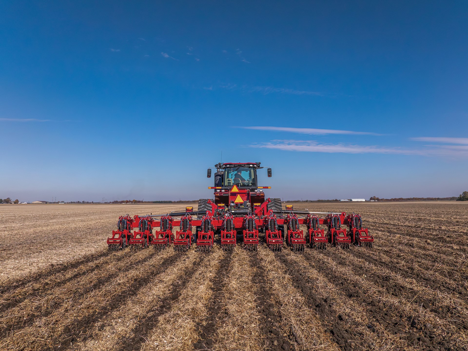 A large red tractor equipped with multiple farming attachments is plowing a vast, dry field under a clear blue sky, creating evenly spaced furrows in the soil.