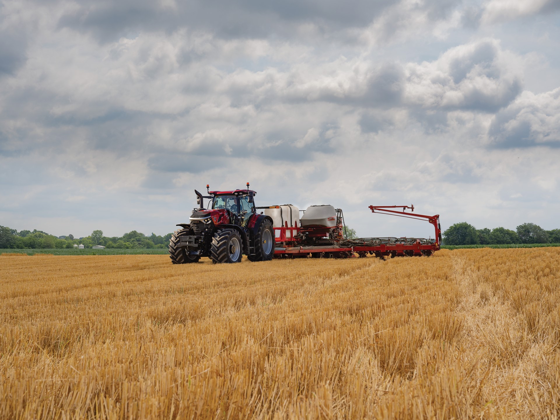 A red tractor with large tires is working in a golden wheat field under a cloudy sky, pulling farming equipment used for planting or fertilizing. Trees and greenery are visible in the distant background.